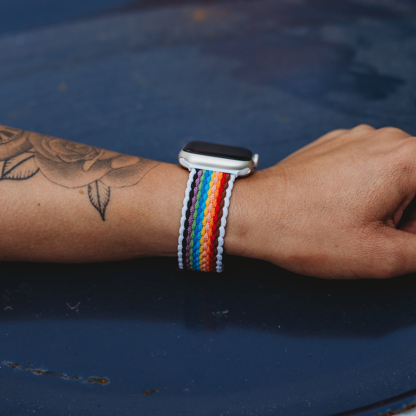 Ein Handgelenk mit Rosentattoo trägt das arktisband Geflochtene Apple Watch Armband "Casual" Rainbow Edition von arktisband, mit bunten Perlen besetzt. Die Hand ruht auf einer blauen Oberfläche.