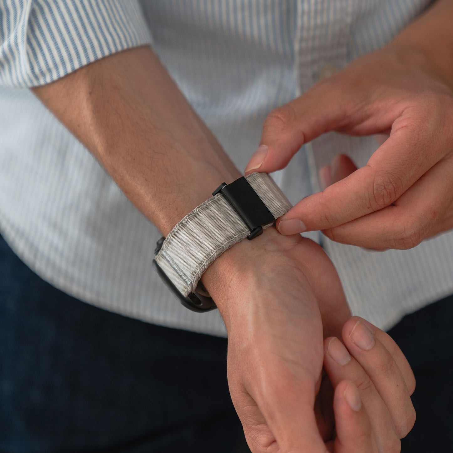 Eine Person befestigt den Verschluss eines Arktisband Apple Watch Nylonarmbands „Venture“ an ihrem Handgelenk und trägt ein hellblau gestreiftes Langarmhemd und eine dunkle Hose.