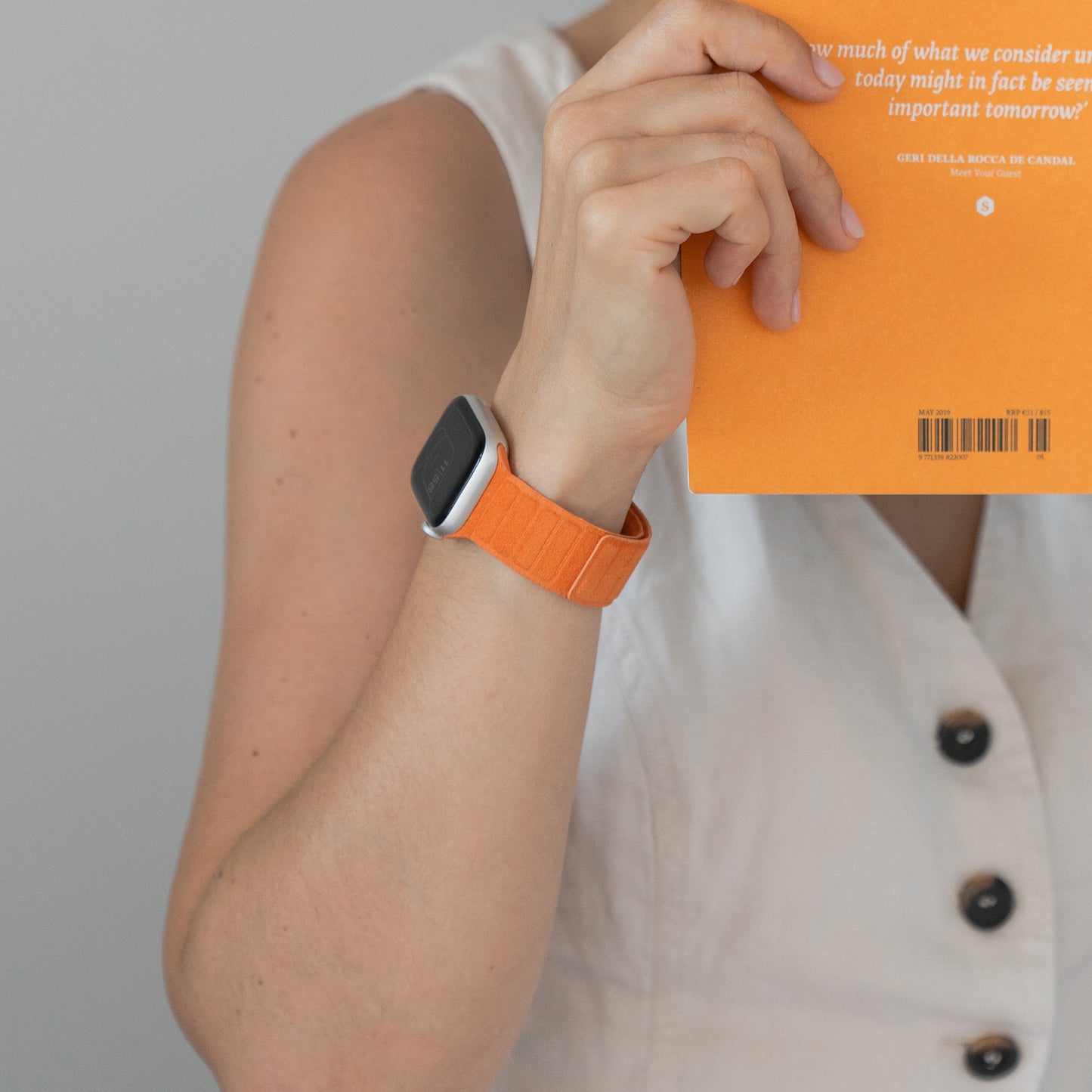 Eine Person in einem weißen, ärmellosen Oberteil hält ein orangefarbenes Buch und trägt das arktisband Apple Watch magnetisches Armband aus Alcantara in orange.