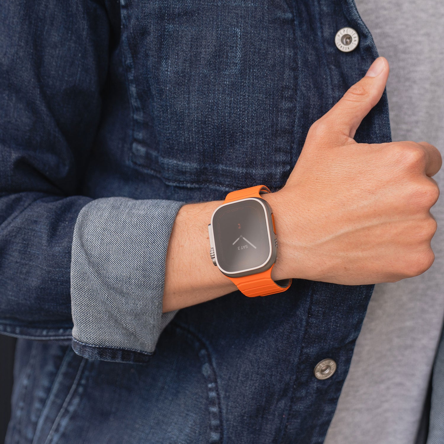 Eine Person in Jeansjacke und einem Apple Watch Magnetarmband „MagFlow“ mit orangefarbenem Band am linken Handgelenk zeigt den Daumen hoch. Die Uhr zeigt ein minimalistisches Zifferblatt.