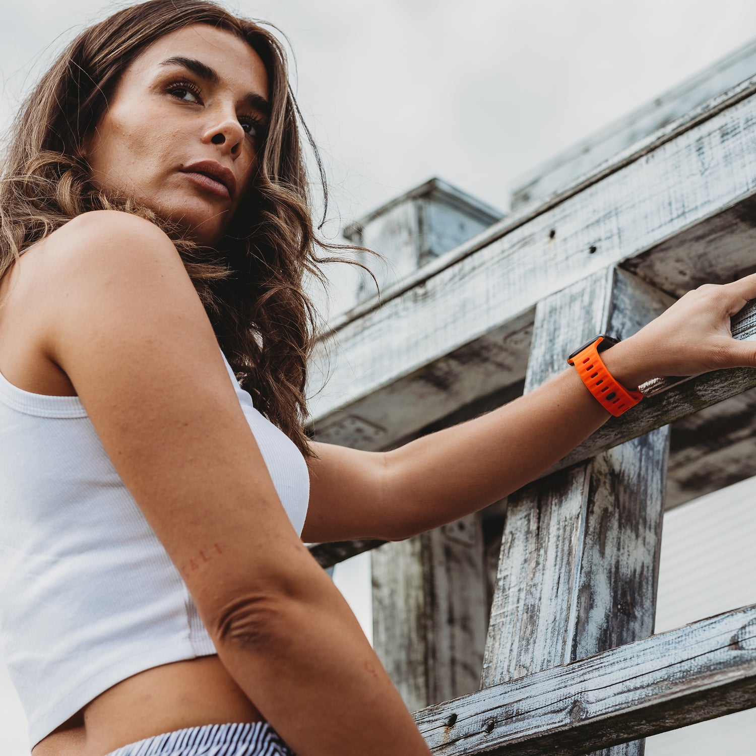Eine Person mit langen braunen Haaren, einem weißen ärmellosen Top und gestreiften Hosen steht im Freien und hält eine verwitterte Holzkonstruktion. Sie trägt ein orangefarbenes Arktisband Apple Watch Silikonarmband „Active“ und blickt leicht nach oben.