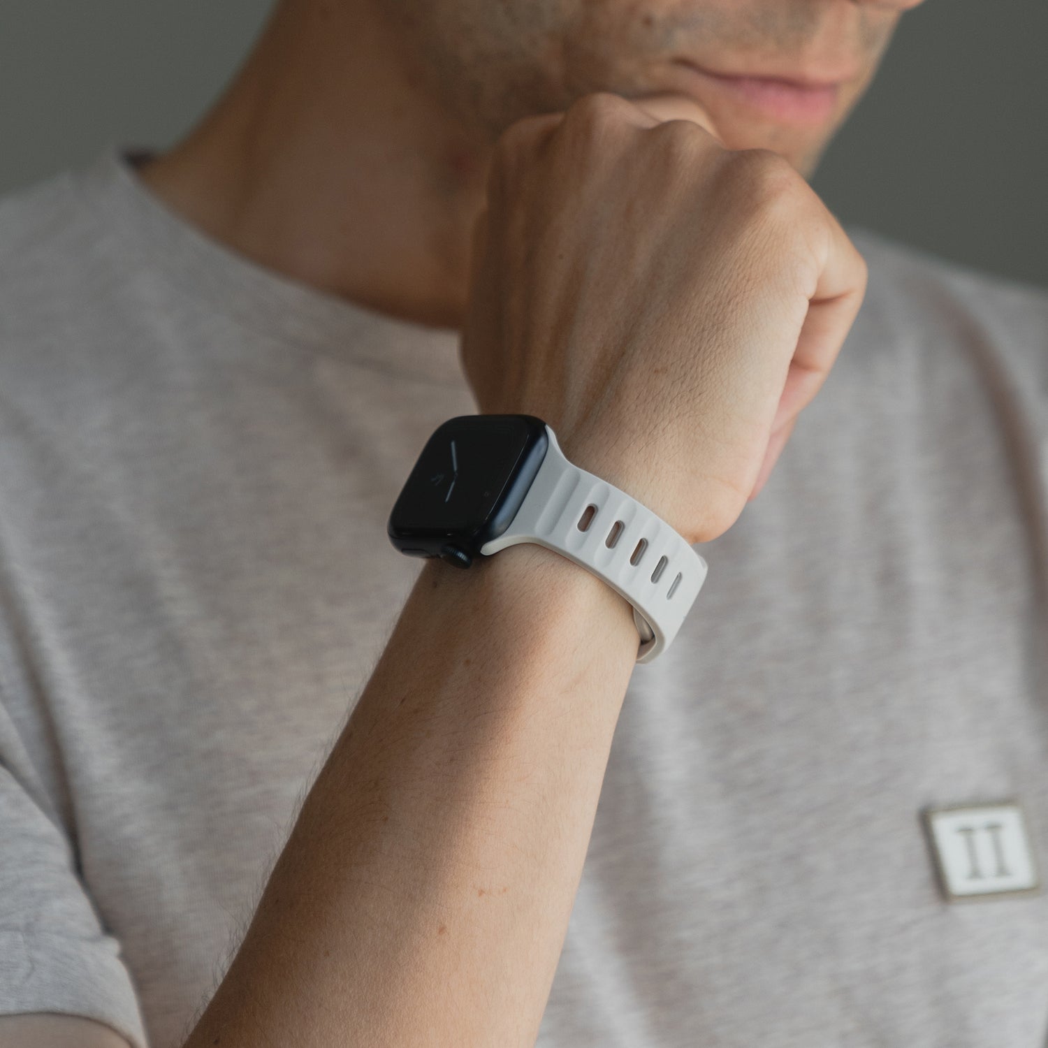 Eine Person im hellgrauen T-Shirt trägt ein arktisband Apple Watch Silikonarmband „Active“ von arktisband in Schwarz mit weißem, atmungsaktivem Silikonband und stützt ihr Kinn auf den angewinkelten Arm; das Zifferblatt ist von der Kamera abgewandt.