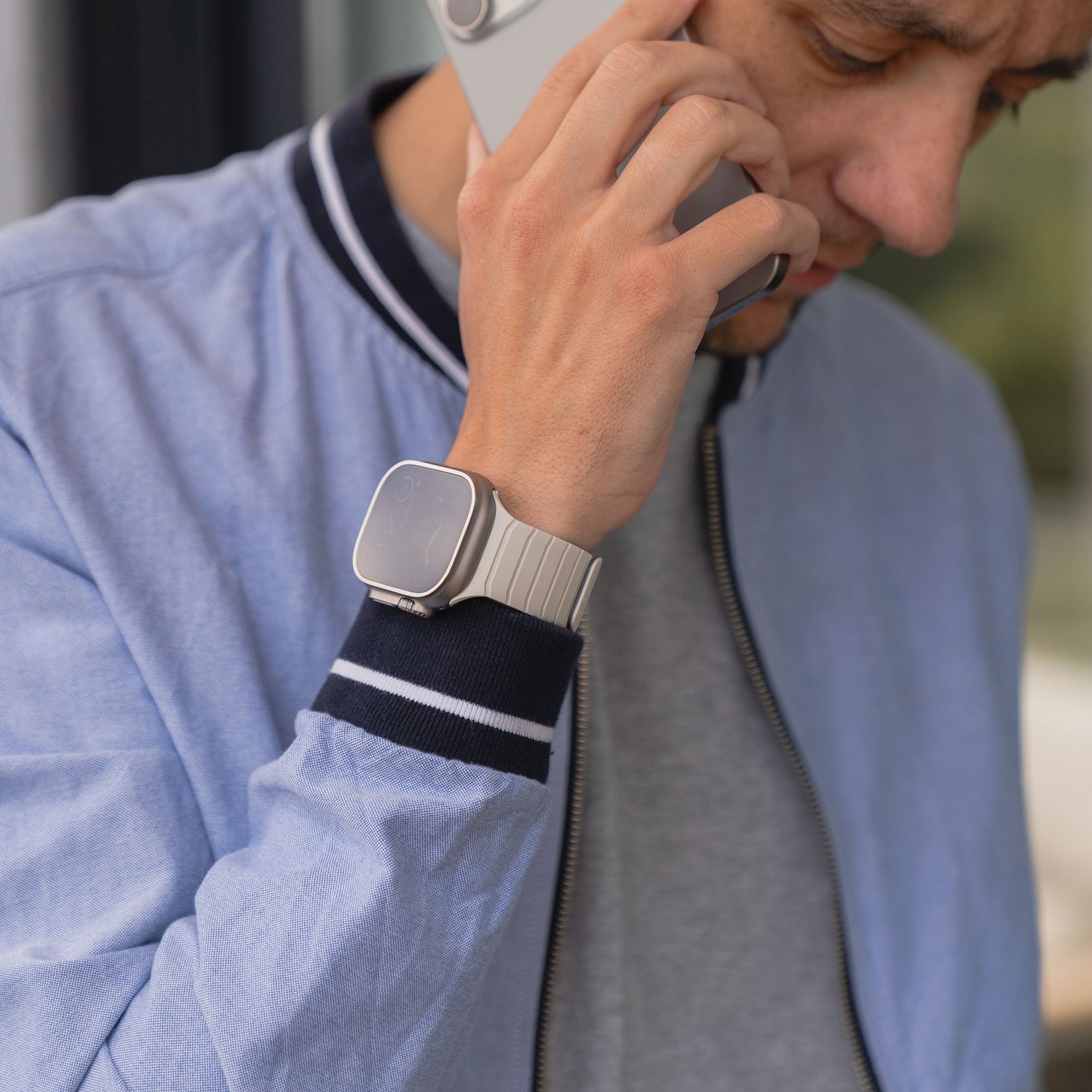 Ein Mann in einer hellblauen Jacke telefoniert mit seinem Smartphone und trägt ein Arktisband Apple Watch Magnetarmband „MagFlow“ aus Silikon mit Magnethalterung am Handgelenk.