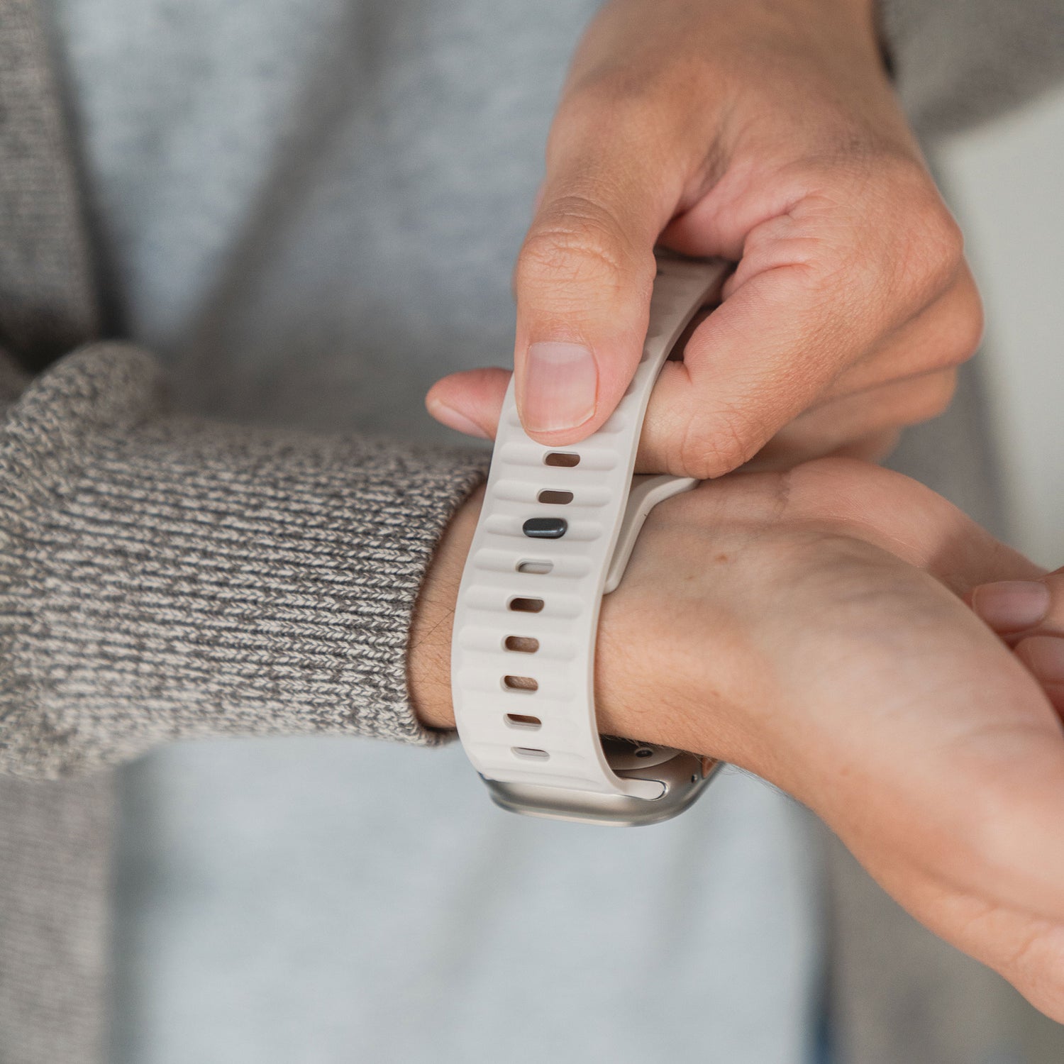 Eine Person befestigt für einen sportlichen Look ein Arktisband Apple Watch Silikonarmband „Active“ in Weiß am Handgelenk, trägt einen grauen Pullover und richtet das Band mit der anderen Hand.