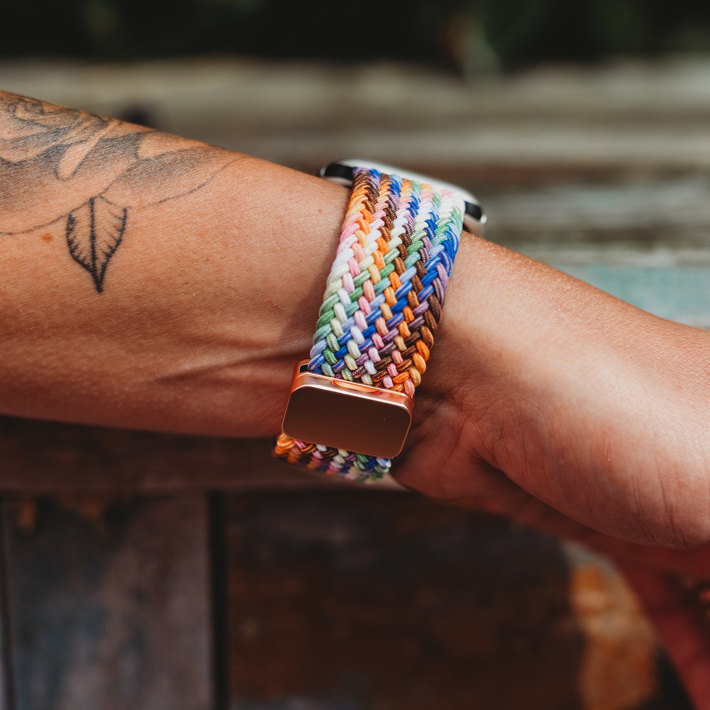 Eine Nahaufnahme zeigt ein Handgelenk mit einem Blatttattoo, das das arktisband Geflochtene Apple Watch Armband „Casual“ Rainbow Edition aus geflochtenem Polyestergarn mit magnetischem Metallverschluss trägt. Der Hintergrund ist leicht unscharf.