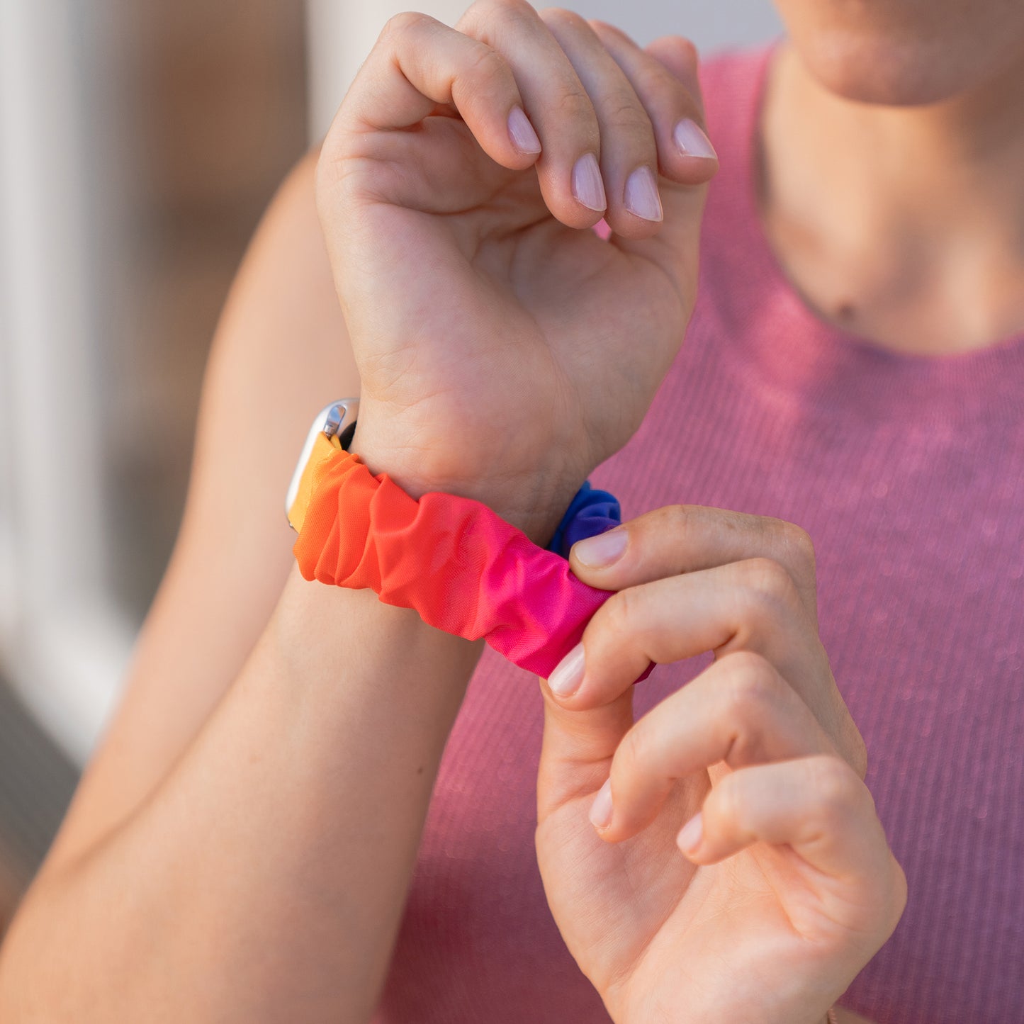 Jemand passt ein arktisband Apple Watch Scrunchie Armband "Elanora" von arktisband mit einem farbenfrohen elastischen Design an, das mit einem rosa ärmellosen Oberteil getragen wird.