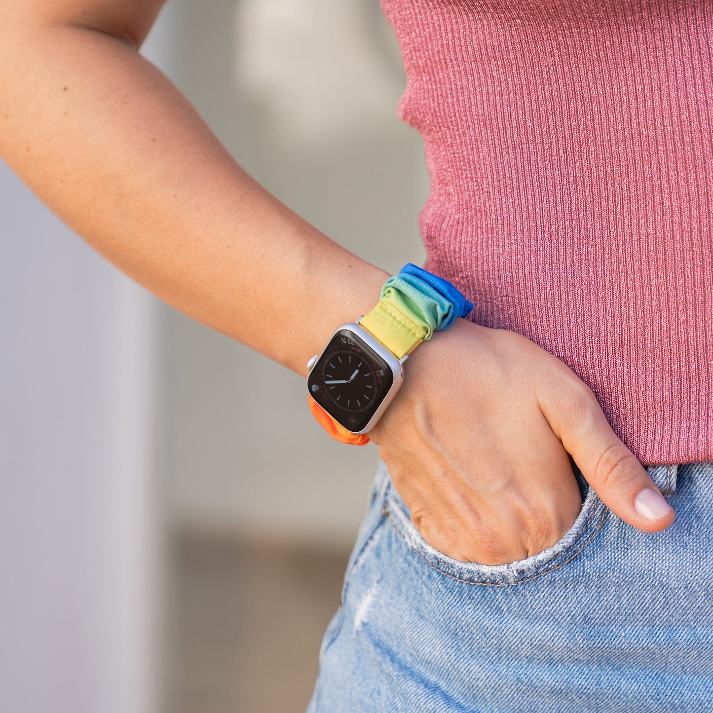 Eine Person trägt ein arktisband Apple Watch Scrunchie Armband "Elanora" - ein farbenfrohes, elastisches Band - am Handgelenk, mit der Hand in der Tasche, bekleidet mit einem pinkfarbenen Oberteil und blauen Jeans.