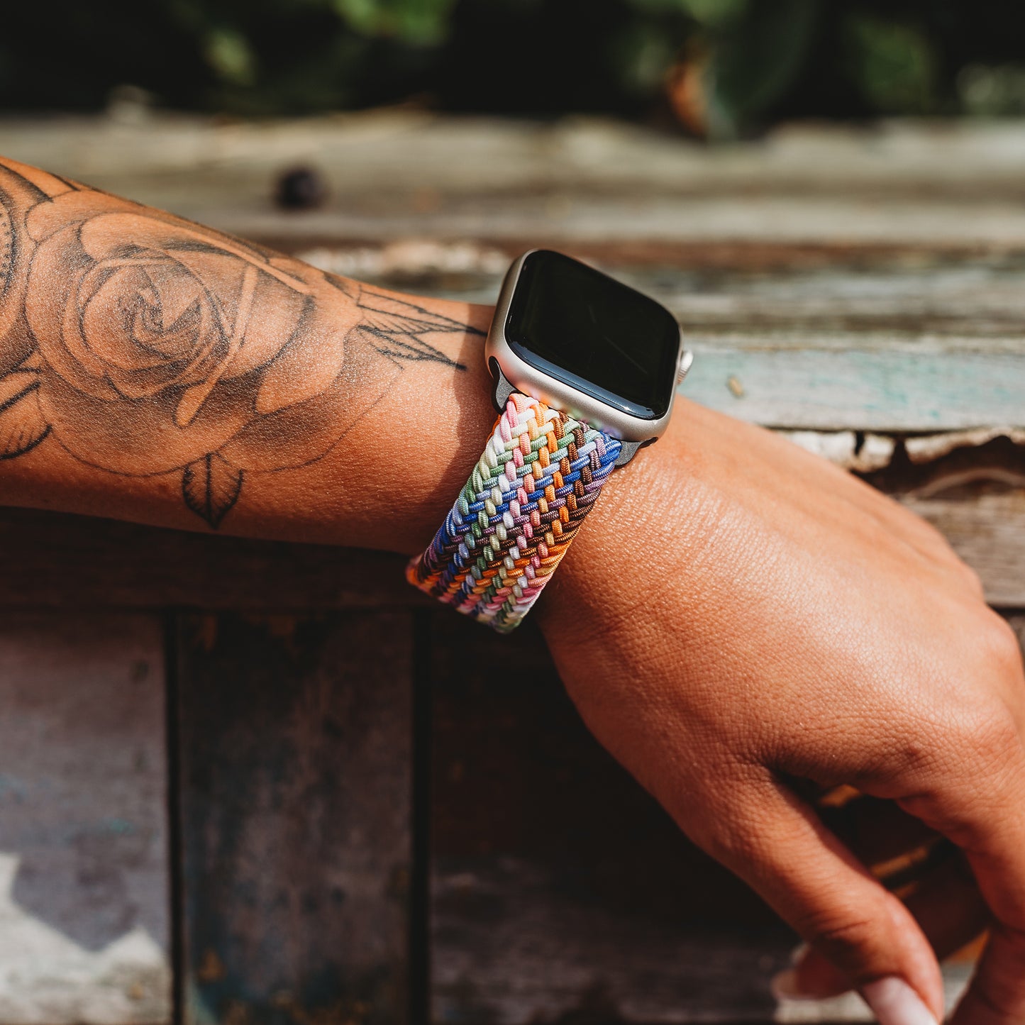 Eine Person mit einem Rosentattoo auf dem Unterarm trägt das arktisband Geflochtene Apple Watch Armband „Casual“ Rainbow Edition; ihre Hand ruht auf einer verwitterten Holzoberfläche mit abgeplatzter Farbe.