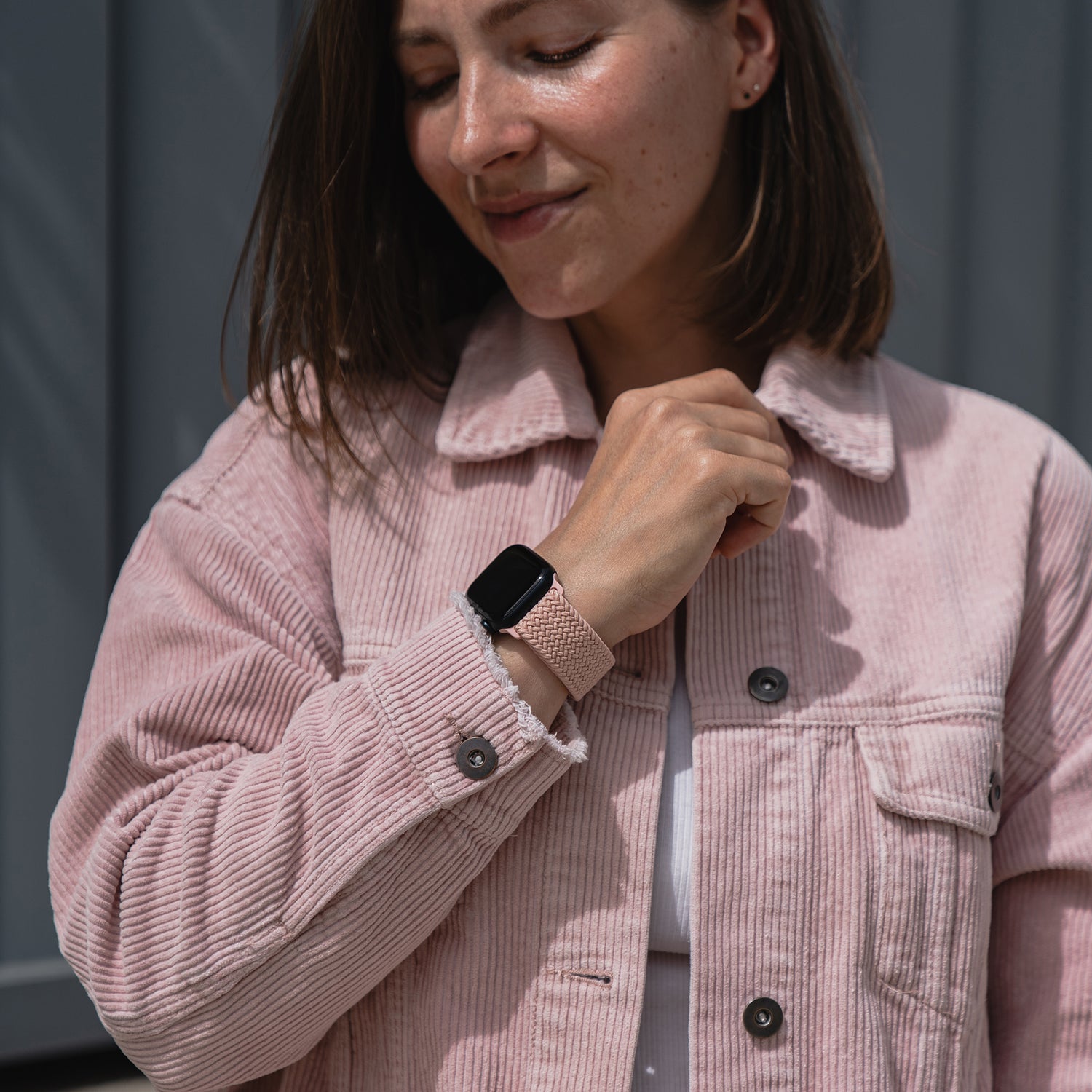 Eine Frau in einer rosa Cordjacke lächelt, während sie im Freien auf ihre Apple Watch schaut, die mit dem geflochtenen Apple Watch Armband „Casual“ mit Magnetverschluss von Arktisband gestylt ist.