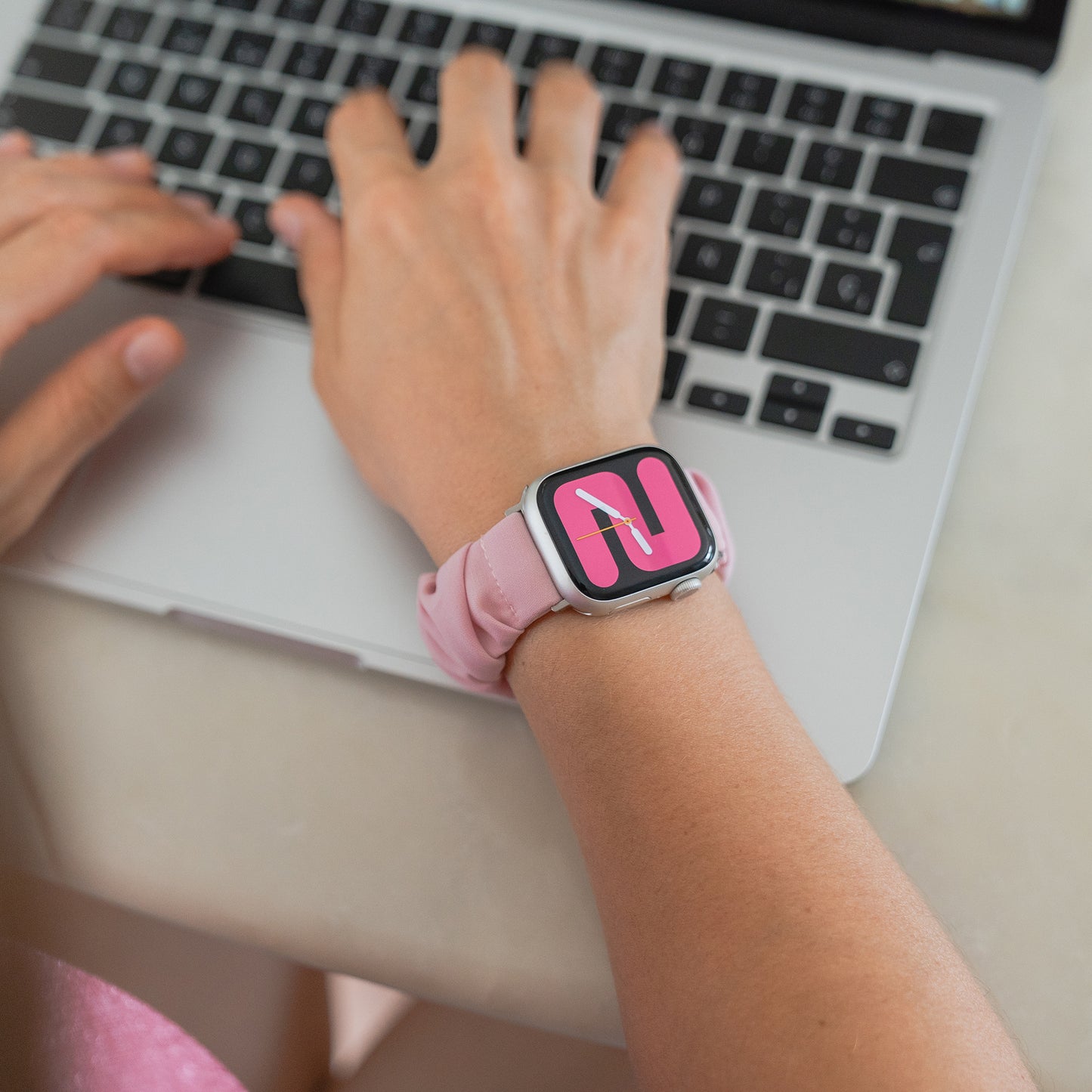 Jemand tippt auf einem Laptop, während er das arktisband Apple Watch Scrunchie Armband "Elanora" trägt, ein rosafarbenes Gummiband von arktisband, gepaart mit einem bunten Zifferblatt.