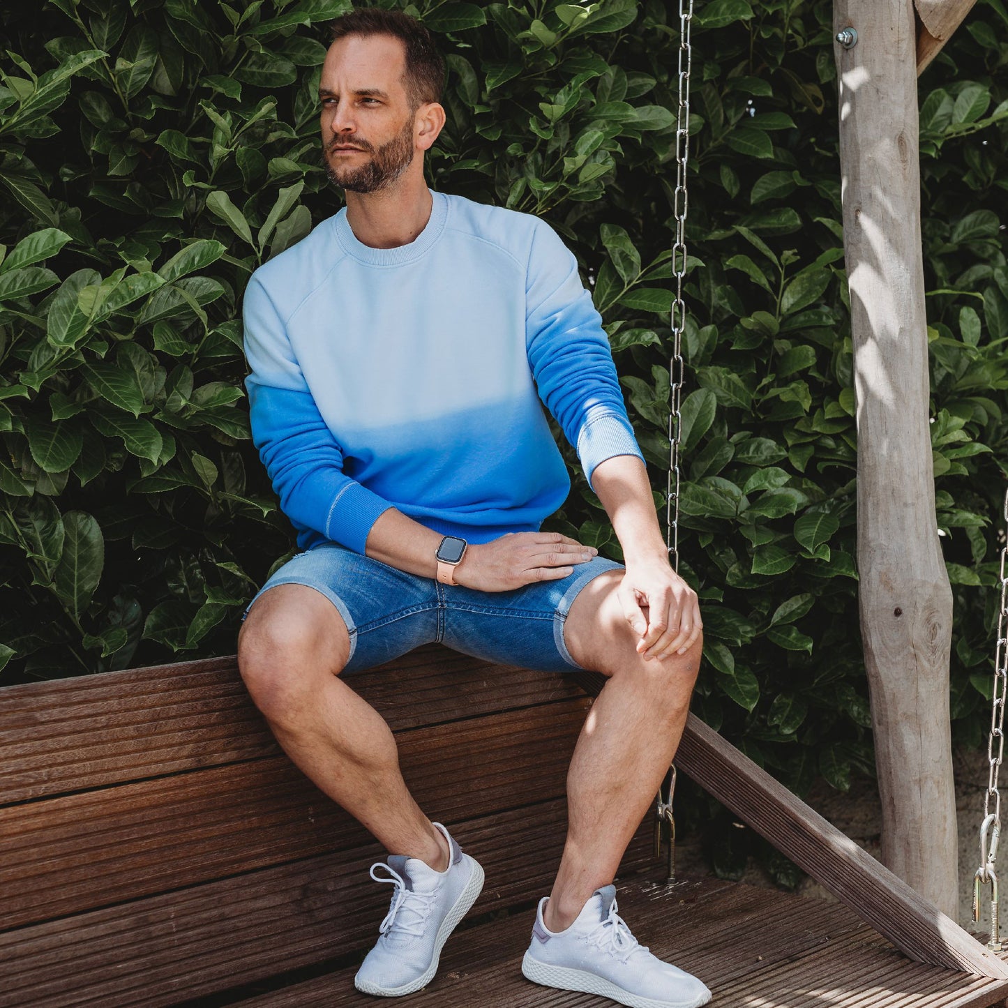 Ein bärtiger Mann sitzt auf einer Holzschaukel. Er trägt ein blaues Sweatshirt mit Farbverlauf, Jeansshorts, weiße Turnschuhe und ein Arktisband Apple Watch FKM Sportarmband „Sweat“. Er blickt zur Seite, ein Arm ruht auf seinem Knie. Hinter ihm ist grünes Laub.