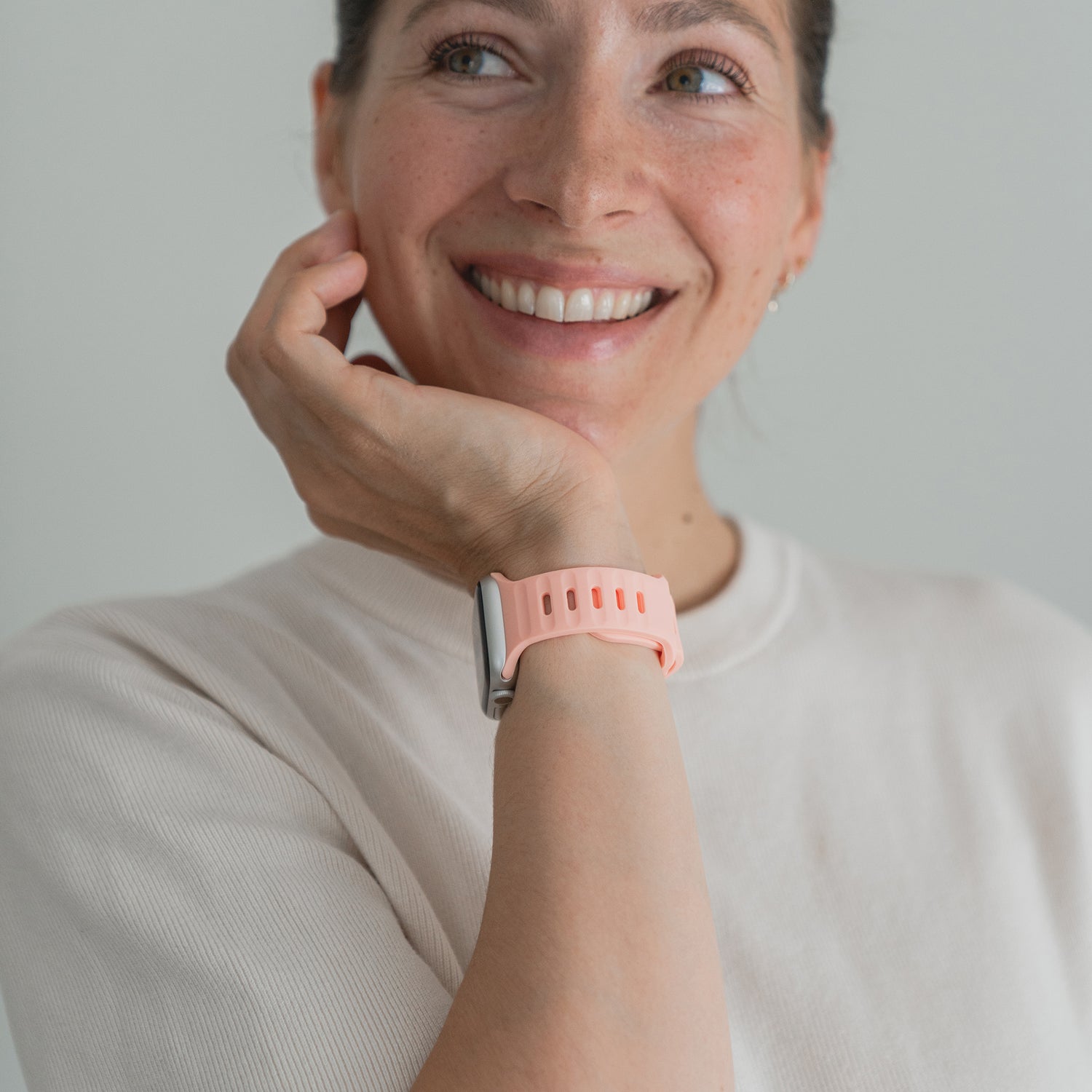Eine Frau in einem hellen Hemd lächelt und stützt ihr Kinn auf ihre Hand. Sie trägt das arktisband Apple Watch Silikonarmband „Active“ mit atmungsaktivem Design und stylischem rosa Band.