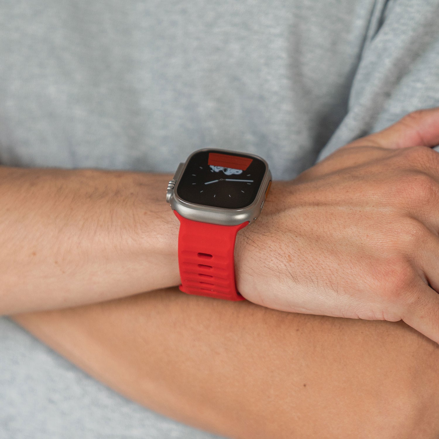 Eine Person im grauen Hemd hat die Arme verschränkt und zeigt ein Apple Watch Silikonarmband „Active“ mit rotem Band am linken Handgelenk. Das atmungsaktive Design der Marke ist teilweise auf dem Display zu sehen.