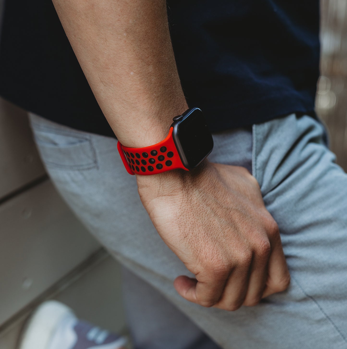 Eine Person in schwarzem Hemd und grauer Hose steht mit einer Hand in der Tasche da und präsentiert das Arktisband Apple Watch Silikonarmband „Energy“ mit rot-schwarzem Lochband an ihrem Handgelenk.