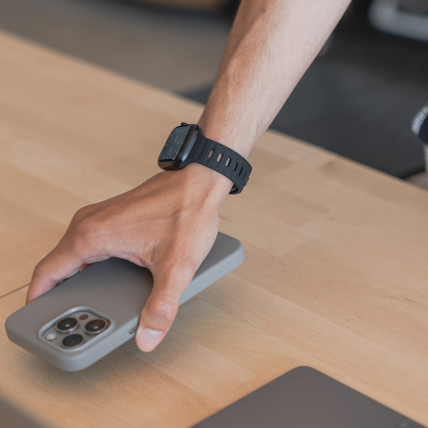 Eine Person, die ein Arktisband Apple Watch Silikonarmband „Active“ mit atmungsaktivem Design trägt, greift nach einem Smartphone mit grauer Hülle auf einem hellen Holztisch.