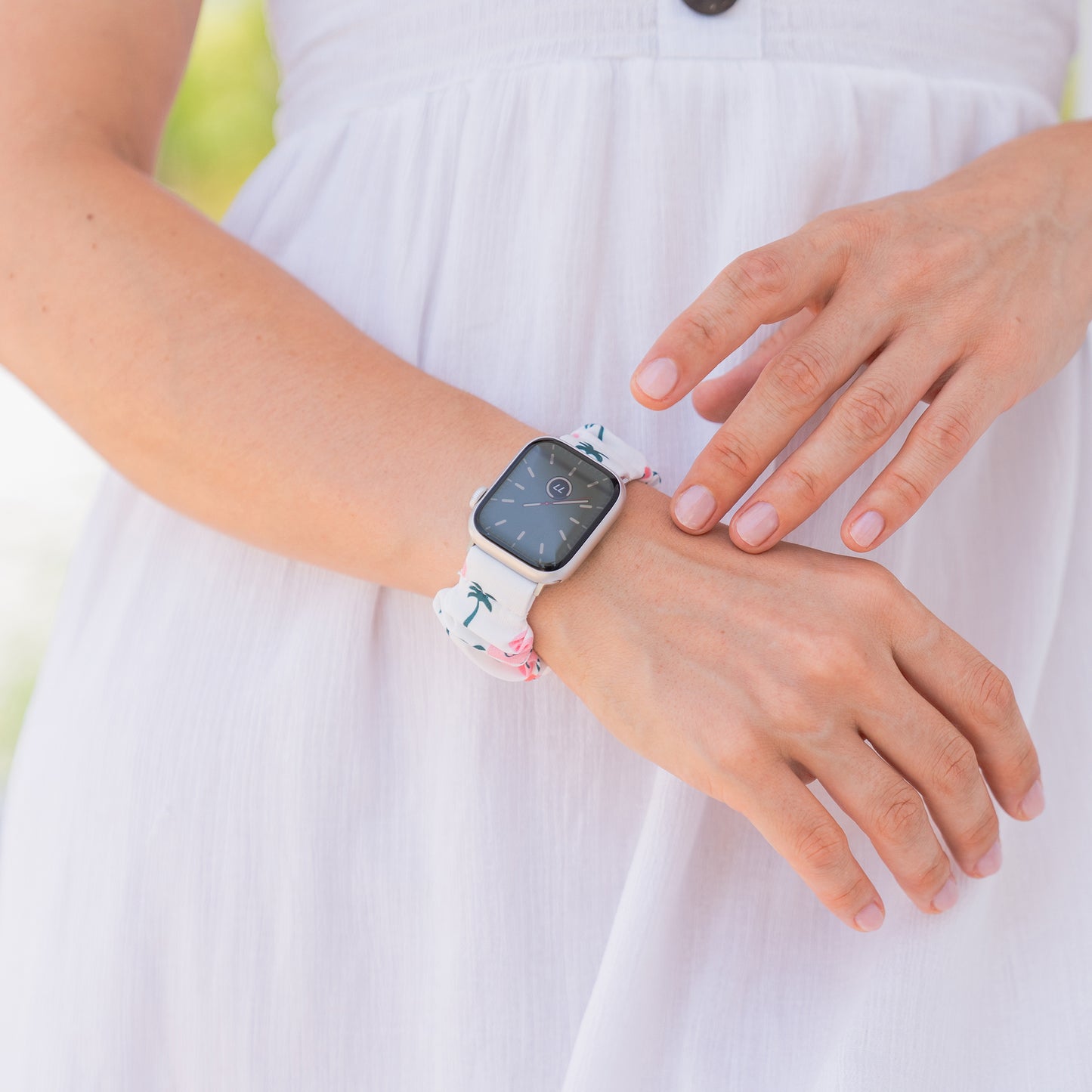Eine Person in einem weißen Kleid trägt ein arktisband Apple Watch Scrunchie Armband "Elanora" mit einem weißen, tropisch gemusterten Band an ihrem Handgelenk.