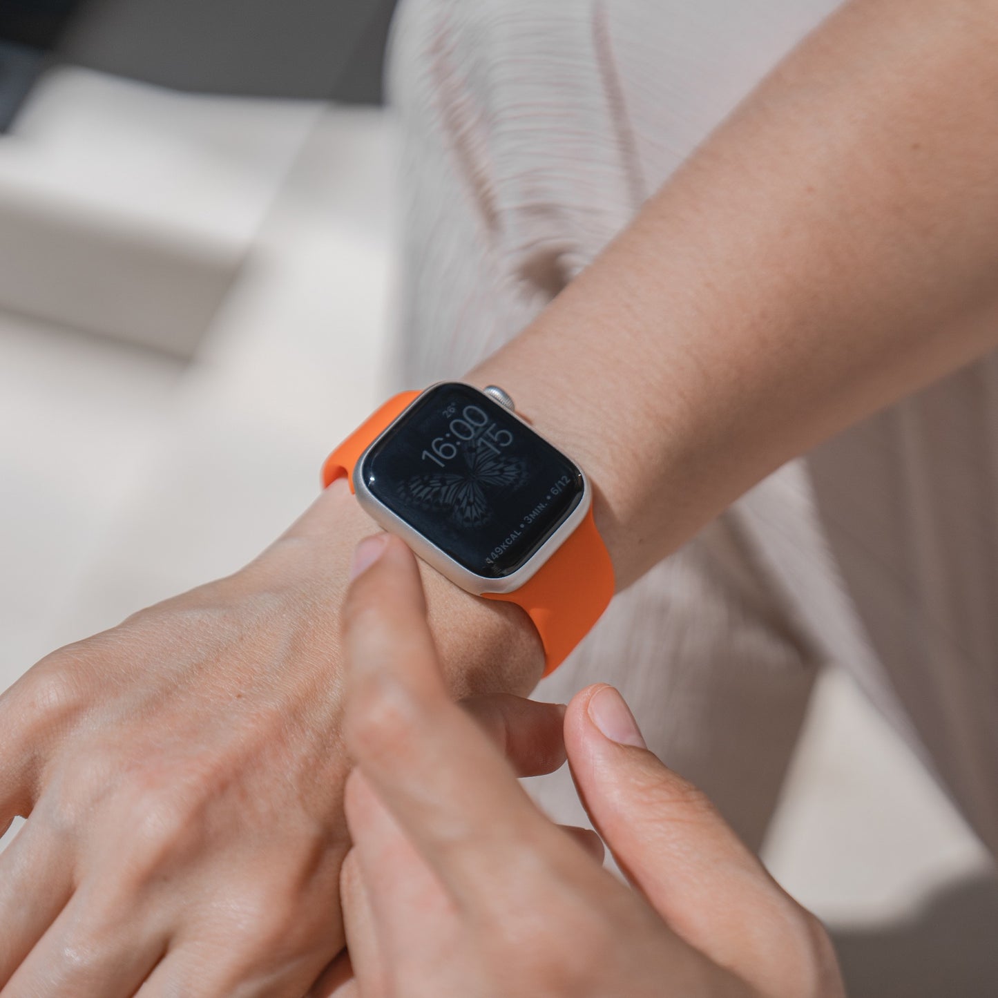 Eine Person trägt das arktisband Apple Watch Silikonarmband „Aqua“ von arktisband mit orangefarbenem Band am linken Handgelenk, schaut auf das Display, das 16:00 und den 15. anzeigt; es ist Apple Watch kompatibel.