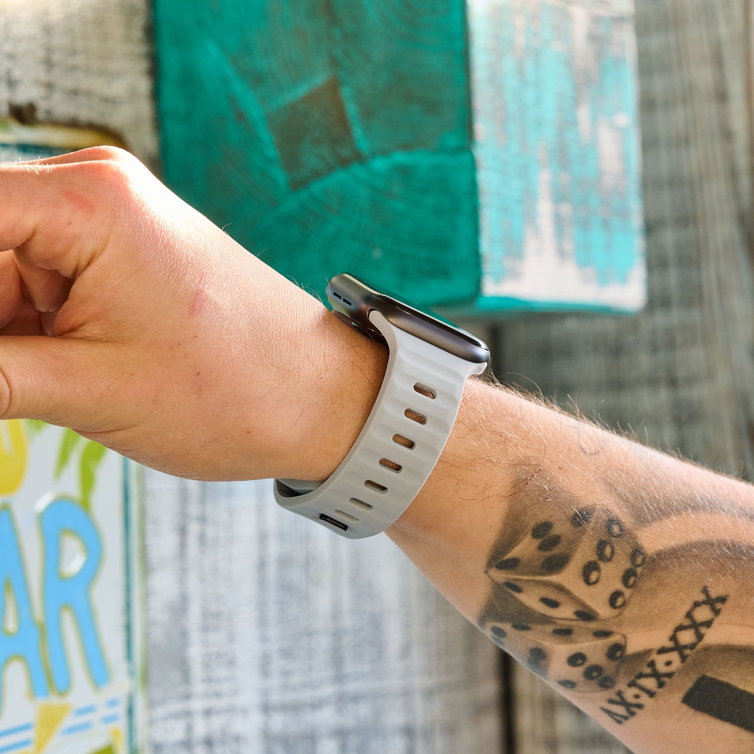 Ein tätowierter linker Arm mit Würfelmotiv trägt ein arktisband Apple Watch Silikonarmband "Active" von arktisband. Im Hintergrund sind Holzbretter und ein teilweise sichtbares gemaltes Schild zu sehen.