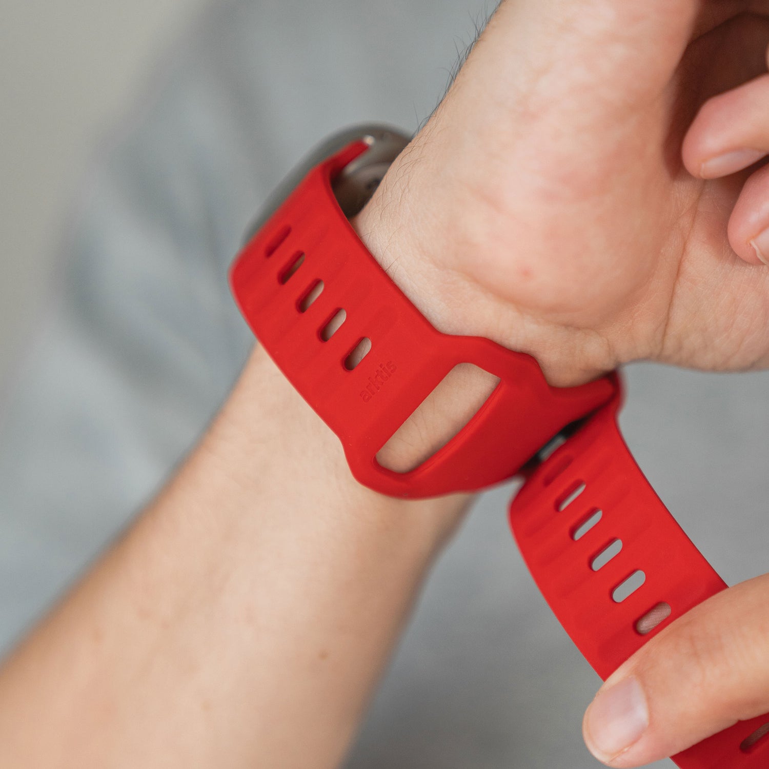 Eine Person befestigt ein Arktisband Apple Watch Silikonarmband „Active“ in leuchtendem Rot am Handgelenk, wobei der Hintergrund leicht unscharf ist.