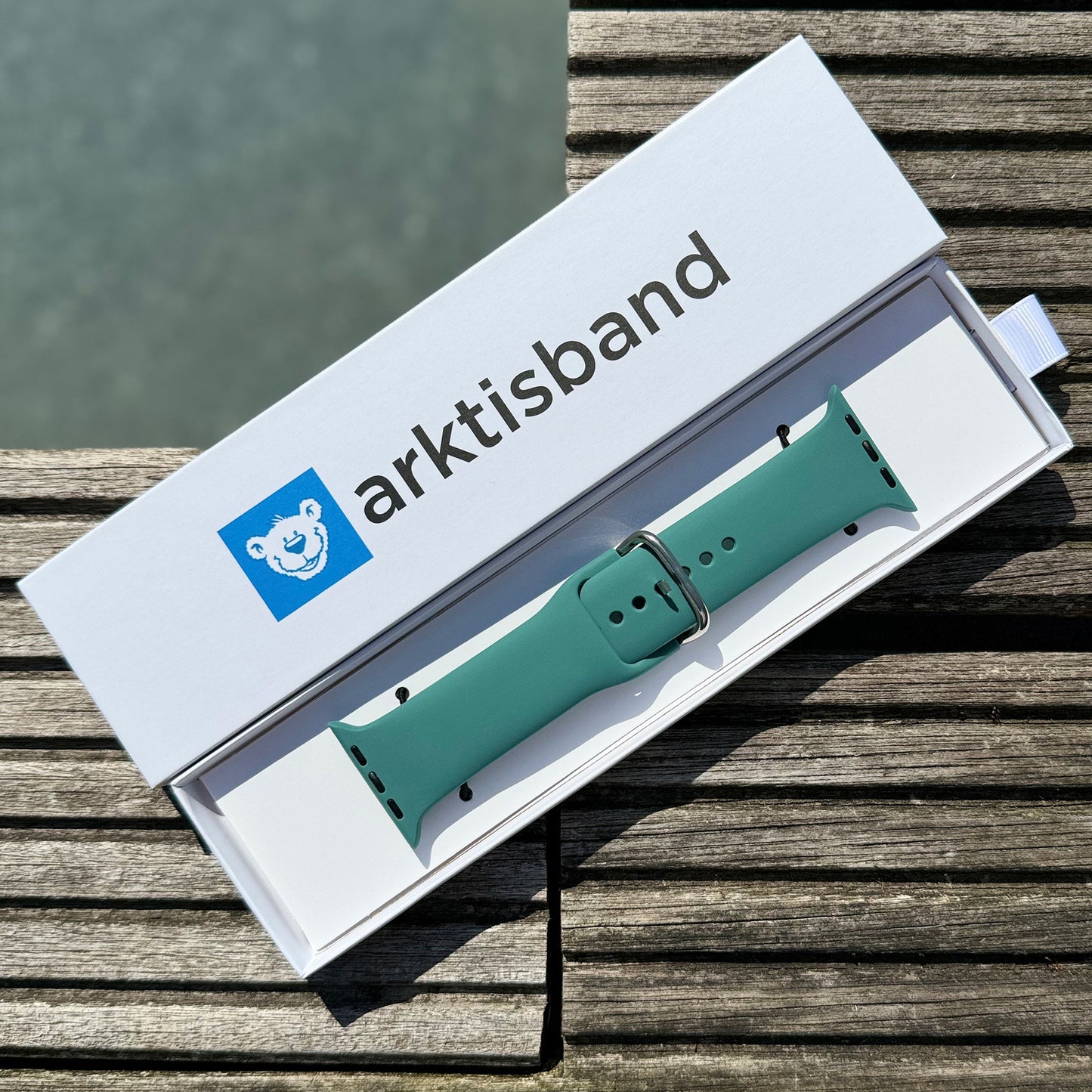 Das arktisband Apple Watch Silikonarmband „Aqua“ mit Metallschließe wird in einer offenen weißen arktisband-Box mit blauem Eisbär-Logo präsentiert, der auf Holz am Wasser ruht – ideal als sportliches Apple Watch-Armband.