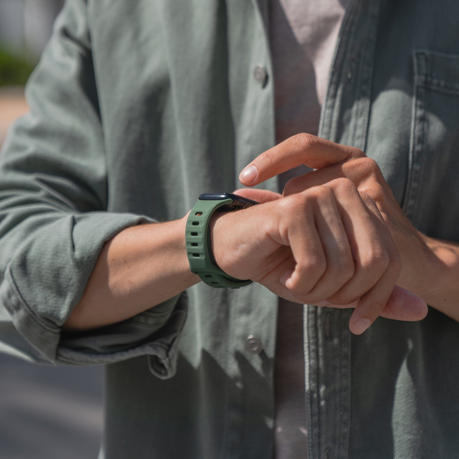 Eine Person tippt im Freien auf das Display eines Arktisband Apple Watch Silikonarmbands „Active“, das sie am linken Handgelenk trägt. Der Fokus liegt auf ihren Händen und der Uhr, ihr Gesicht ist nicht zu sehen.