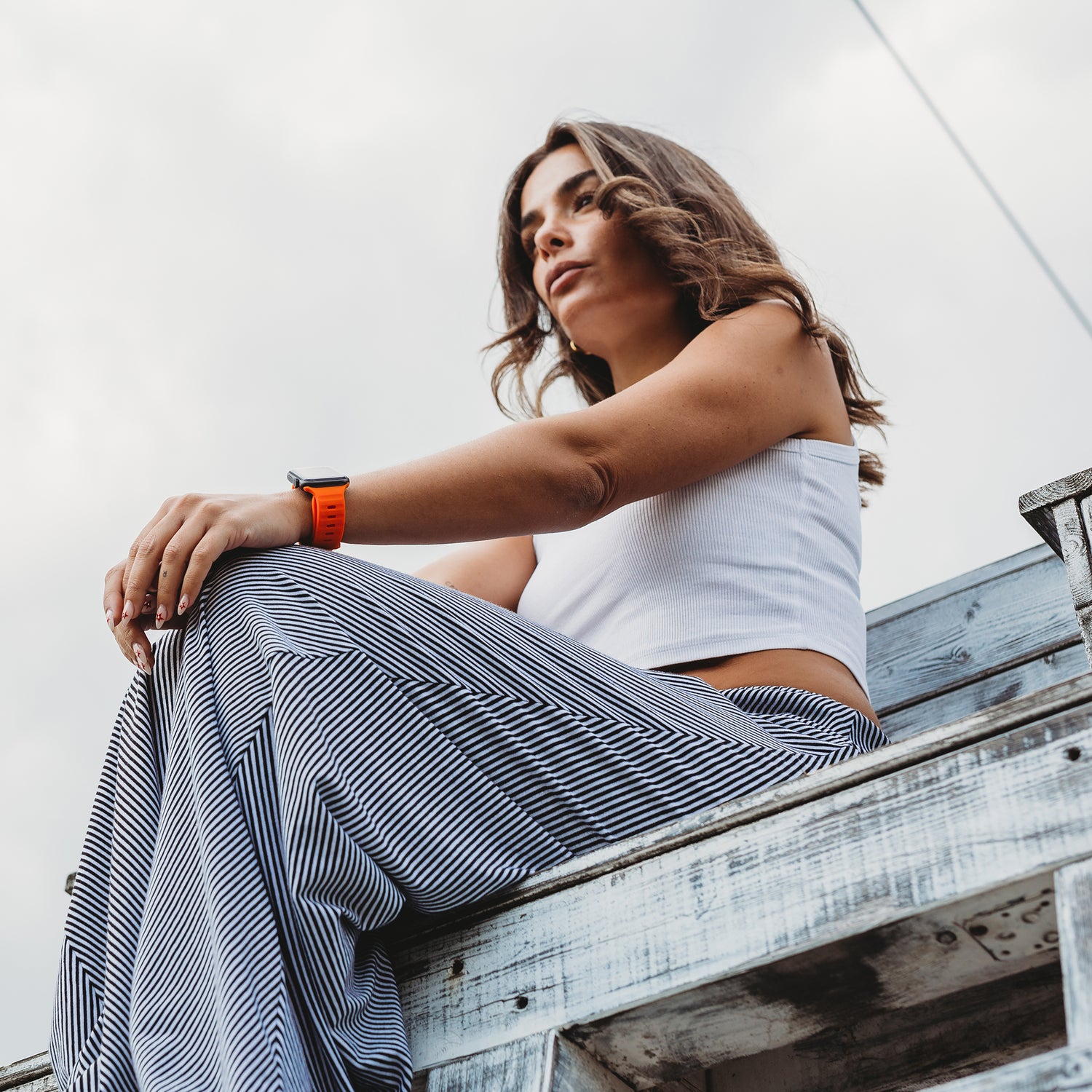 Eine Frau in einem weißen ärmellosen Top und gestreiften Hosen sitzt auf einer verwitterten Holzkonstruktion und blickt in die Ferne. Sie trägt das arktisband Apple Watch Silikonarmband „Active“ von arktisband, und ihr welliges Haar fängt das Licht unter einem bewölkten Himmel ein.