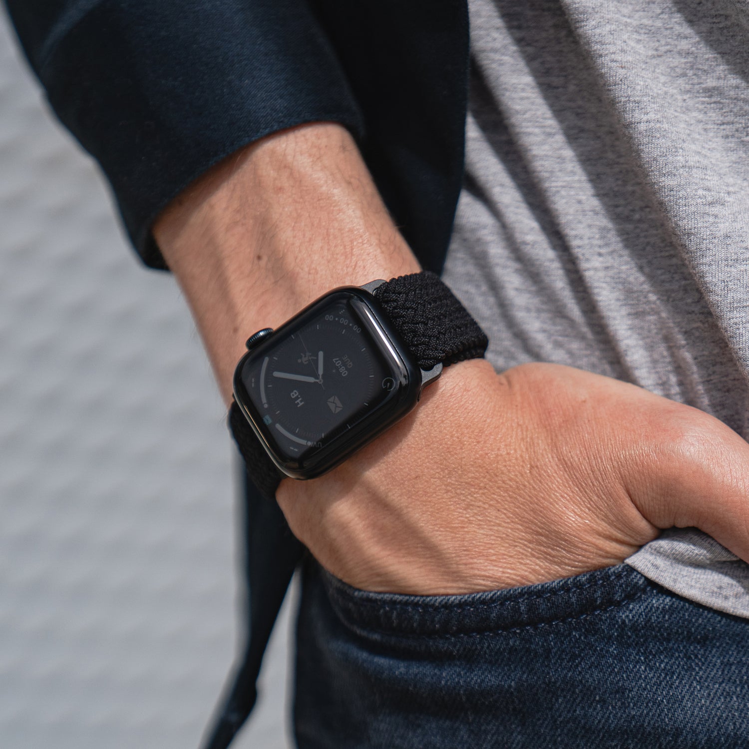 Nahaufnahme des Handgelenks einer Person, die das arktisband Geflochtene Apple Watch Armband „Casual“ mit Magnetverschluss in Schwarz trägt, die Hand in der Tasche hat und leger gekleidet ist.