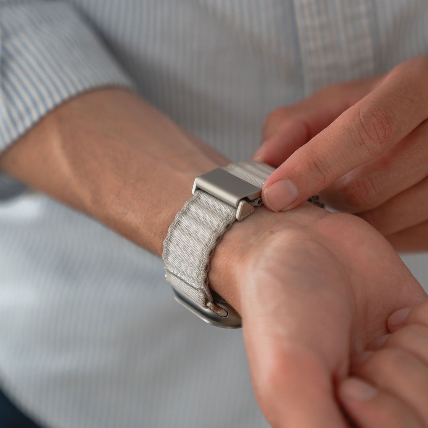 Eine Person im hellblau gestreiften Shirt befestigt ein arktisband Apple Watch Nylon Armband „Venture“ mit Metallschließe am Handgelenk – eine stylische Wahl für Outdoor-Fans, die den perfekten Wellen-Look anstreben.
