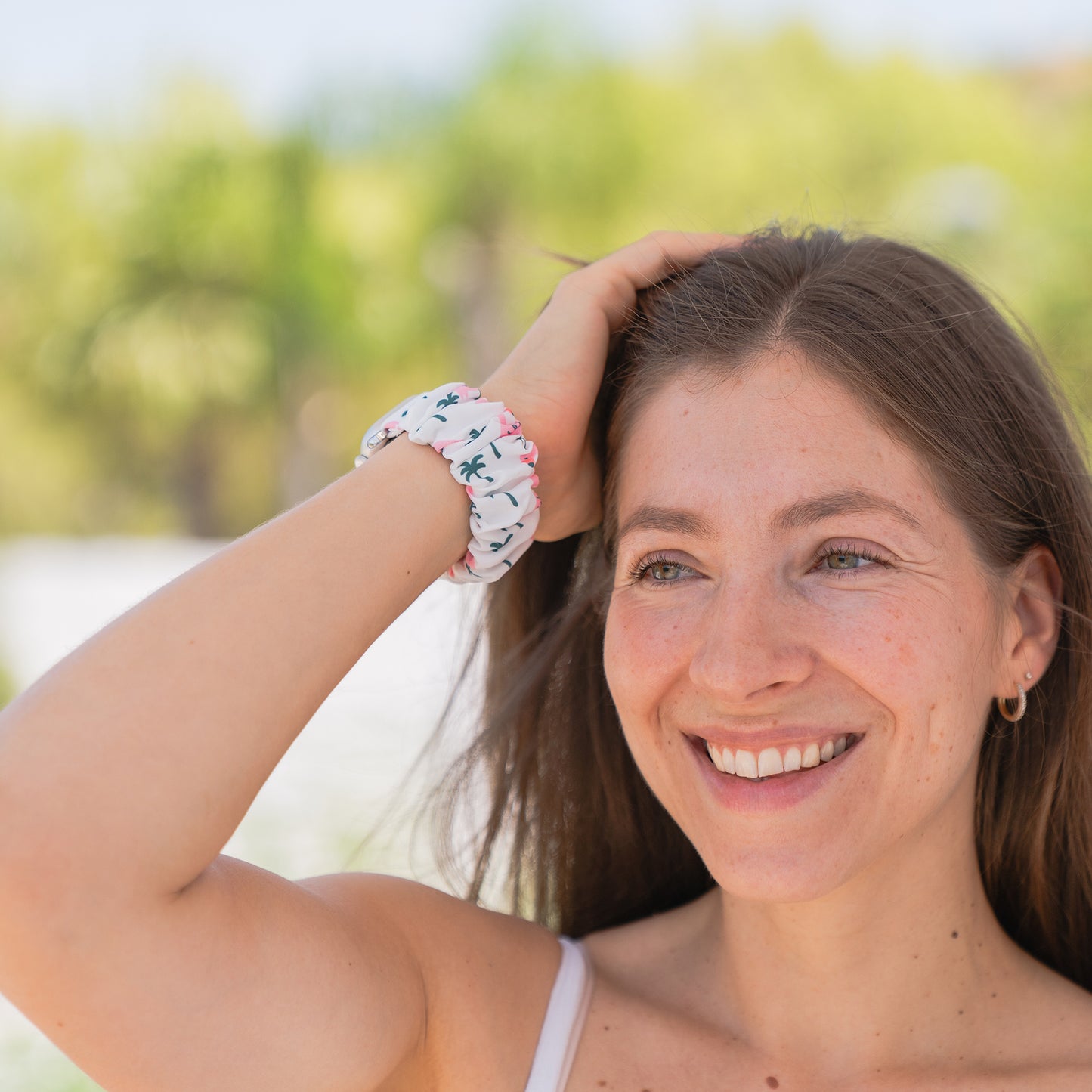 Lächelnde Frau im Freien mit langen braunen Haaren, die das arktisband Apple Watch Scrunchie Armband "Elanora" von arktisband mit einem Palmenmuster trägt.