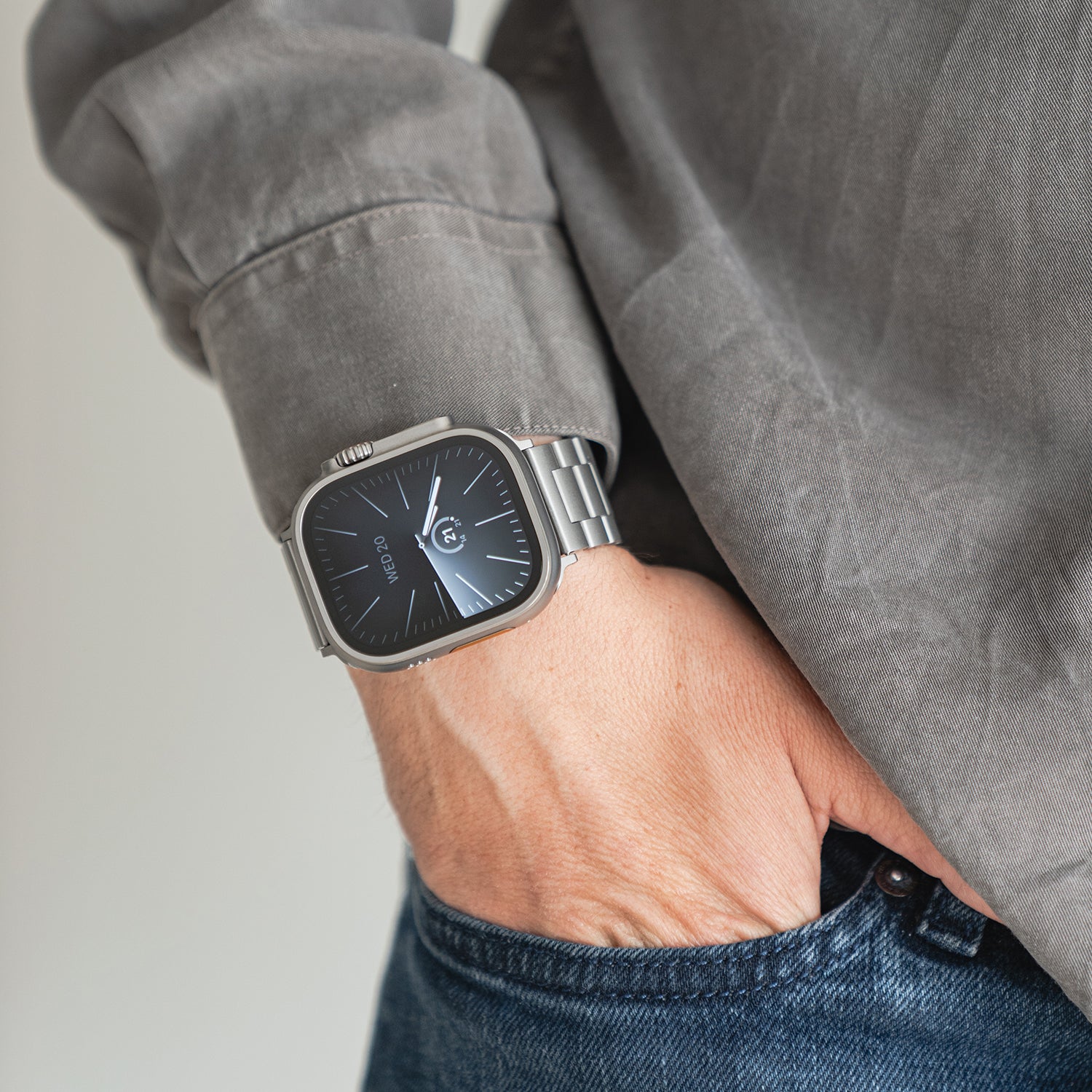 Eine Person in grauem Hemd und blauer Jeans hat die linke Hand in der Hosentasche und zeigt ein Arktisband Apple Watch Gliederarmband „Arctica“ mit schwarzem quadratischem Ziffernblatt am Handgelenk.