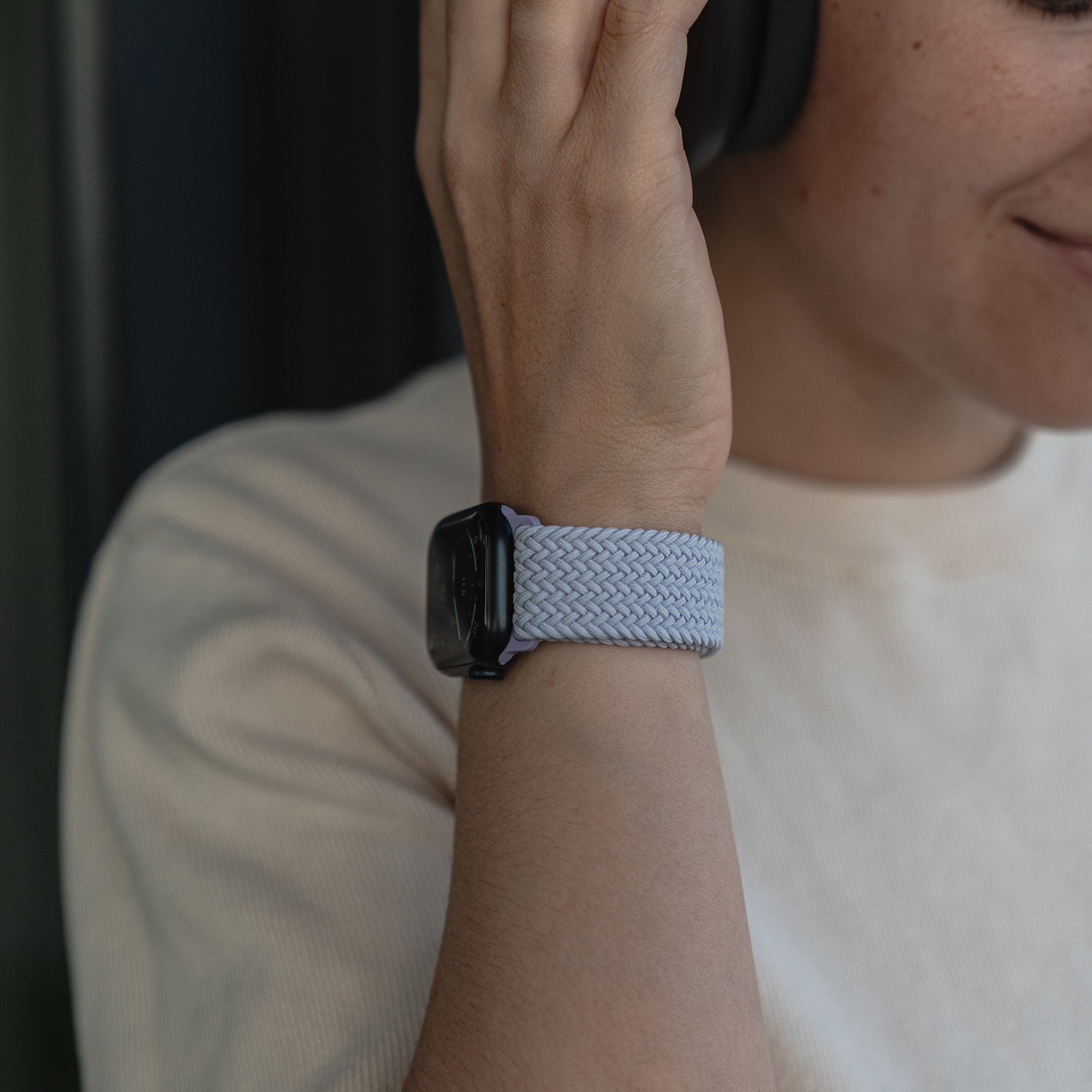 Eine Person trägt das arktisband Geflochtene Apple Watch Armband „Casual“ mit Magnetverschluss in Hellblau, dazu Kopfhörer und ein Hemd in neutraler Farbe und lächelt leicht.