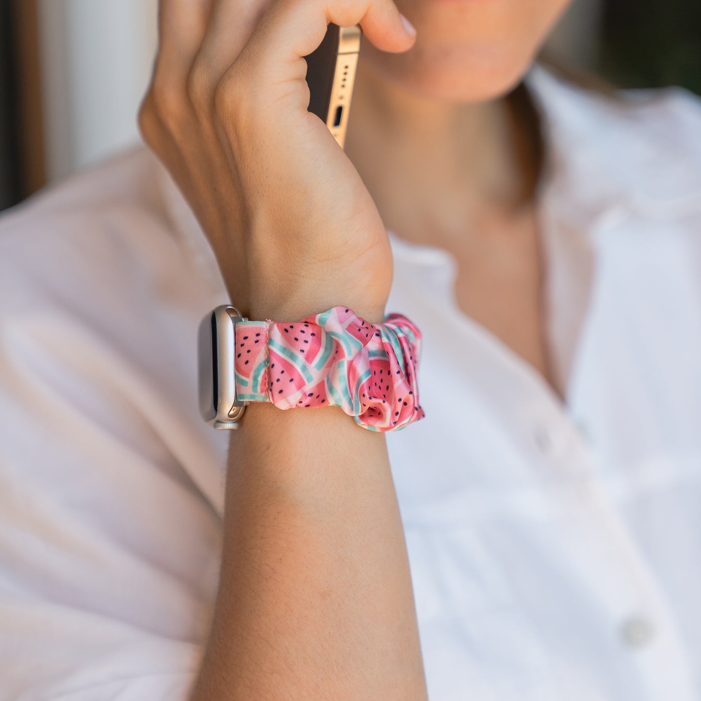 Eine Person in einem weißen Hemd hält ein Telefon, während sie ein Arktisband Apple Watch Scrunchie Armband "Elanora" mit einem Wassermelonenmuster und eine Apple Watch am Handgelenk trägt.