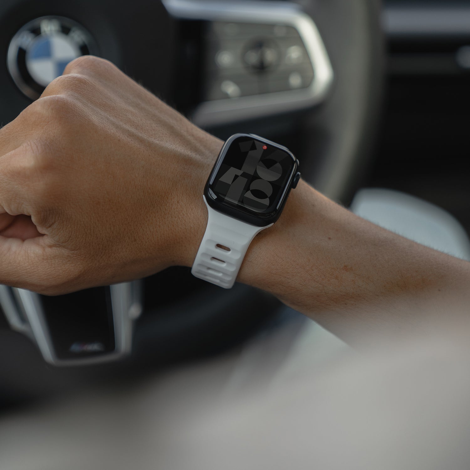 Eine Person mit einem Arktisband Apple Watch Silikonarmband „Active“ in Weiß sitzt in einem Auto und hält das Lenkrad, im Hintergrund ist ein verschwommenes BMW-Logo zu sehen.