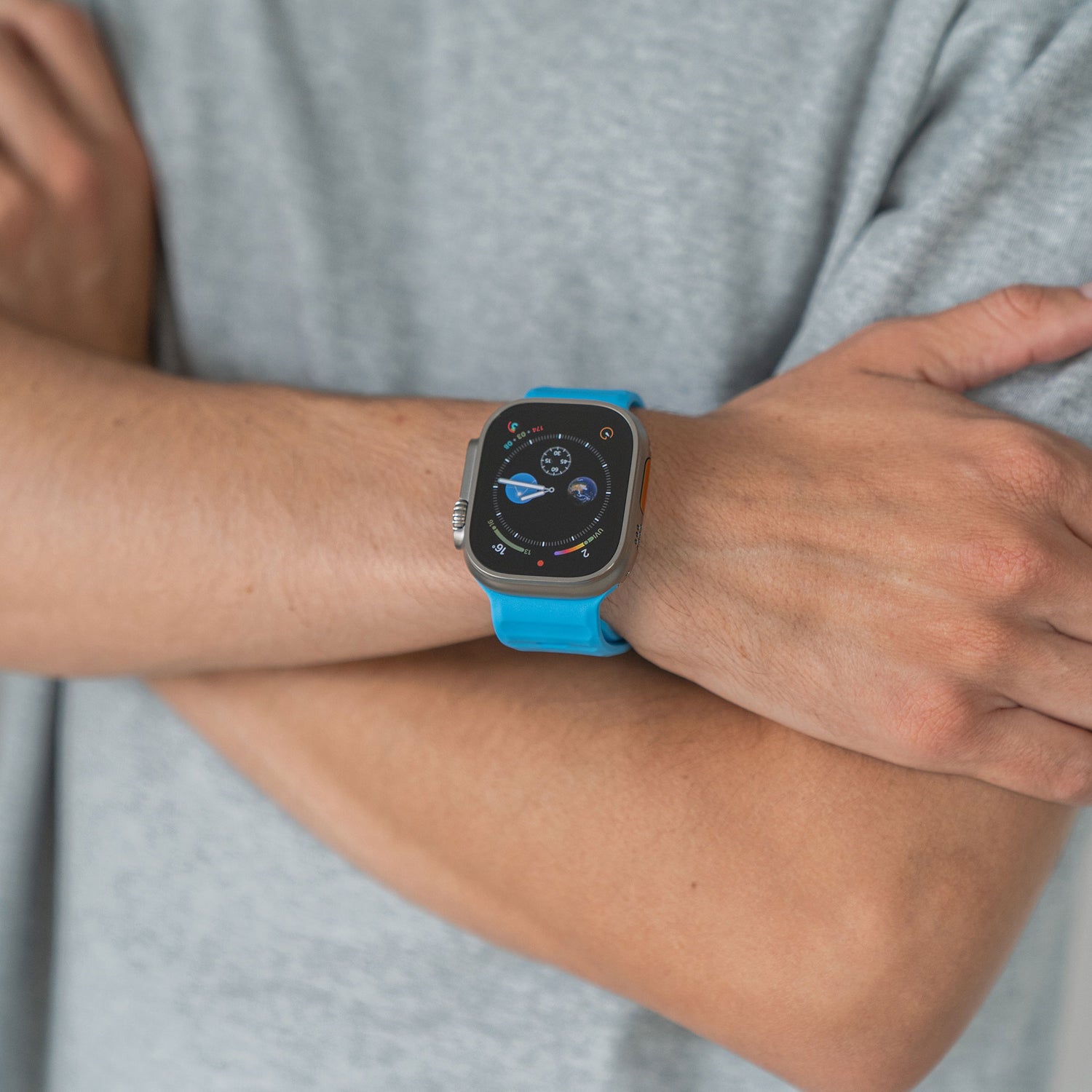 Eine Person in einem hellgrauen Kurzarmhemd verschränkt die Arme und zeigt eine Apple Watch mit einem blauen Silikonarmband „Active“ von arktisband und einer Digitalanzeige am Handgelenk.