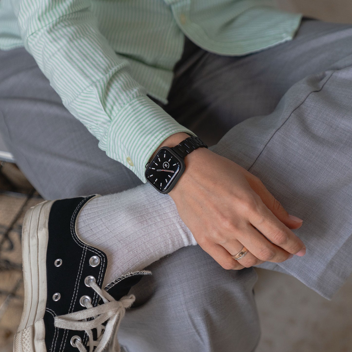 Eine Person in grauer Hose, hellgrünem gestreiftem Hemd und schwarz-weißen Turnschuhen sitzt im Schneidersitz und zeigt an der linken Hand eine Apple Watch mit dem Arktisband „Arctica“ Slim Gliederarmband und einem goldenen Ring.