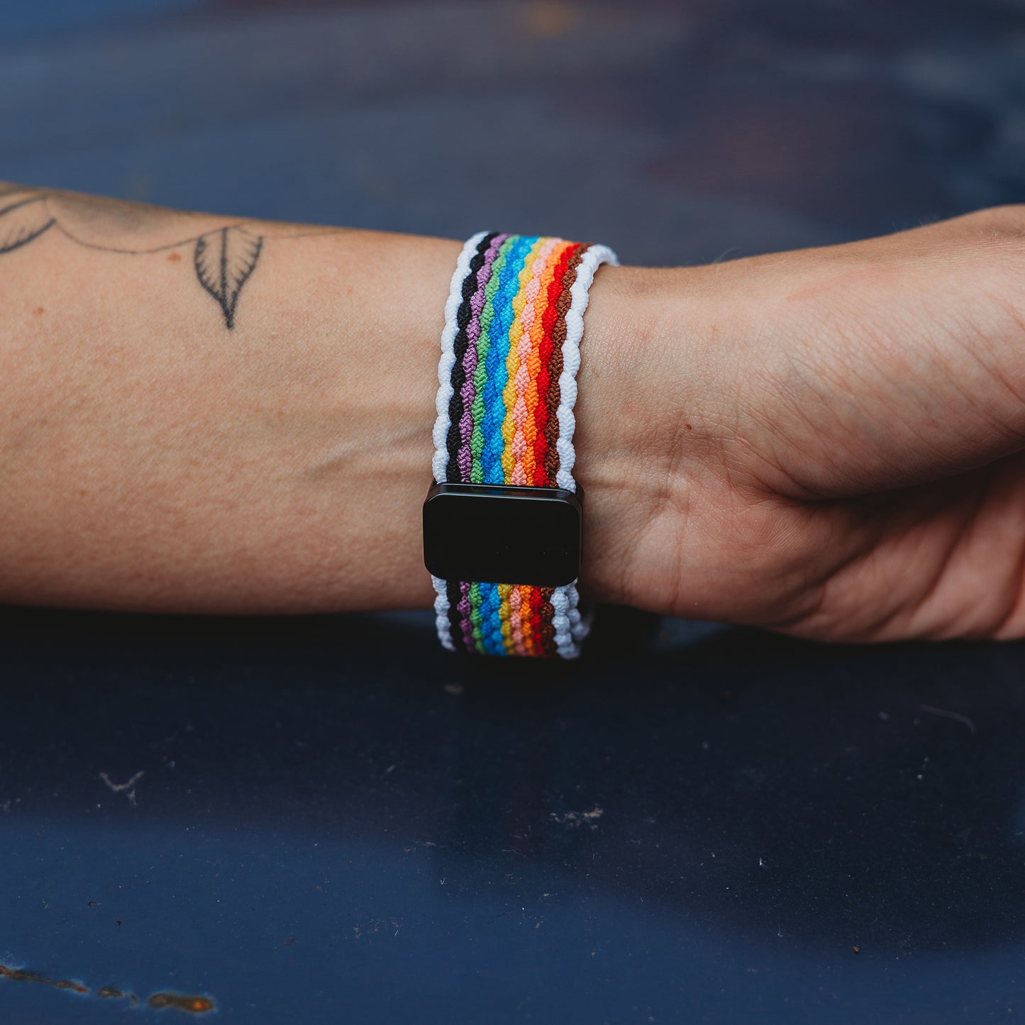 Ein tätowiertes Handgelenk trägt das arktisband Geflochtene Apple Watch Armband „Casual“ Rainbow Edition von arktisband, abgebildet auf einem dunkelblauen Untergrund.