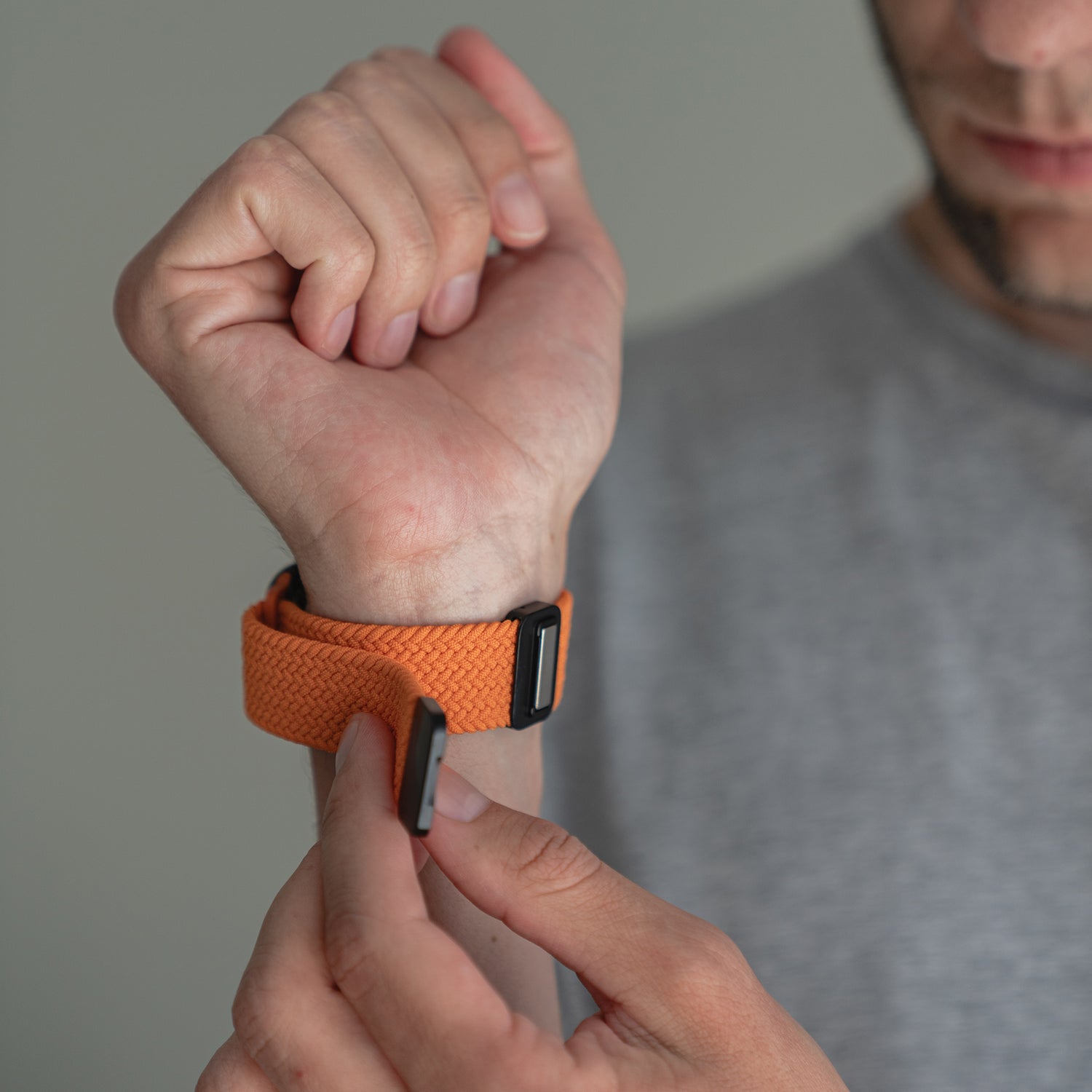 Jemand befestigt ein Arktisband Geflochtenes Apple Watch Armband „Casual“ mit Magnetverschluss in Orange an seinem Handgelenk und trägt dabei ein graues T-Shirt.