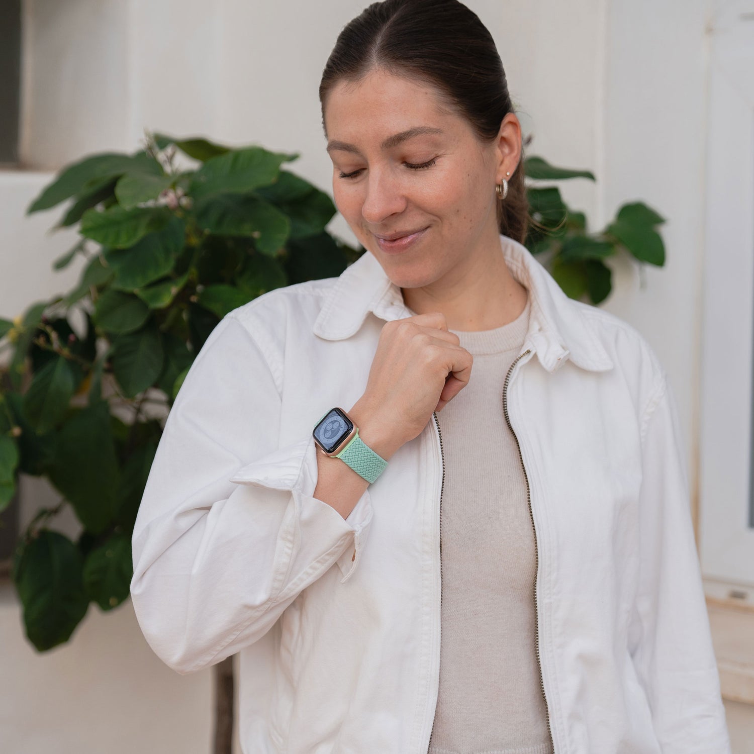 Eine Frau in einer weißen Jacke lächelt, während sie neben einer grünen Blattpflanze steht und ihr geflochtenes Apple Watch Armband „Casual“ mit Magnetverschluss von arktisband überprüft.