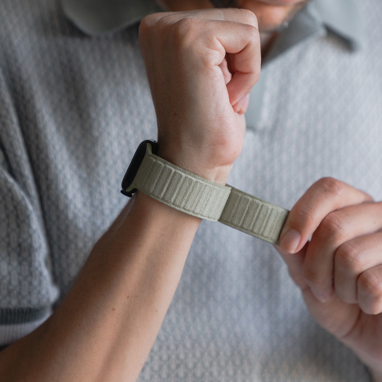 Eine Person befestigt ein arktisband Apple Watch magnetisches Armband aus Alcantara von arktisband, das für seine Langlebigkeit und Pflegeleichtigkeit bekannt ist, während sie ein gemustertes Poloshirt trägt.