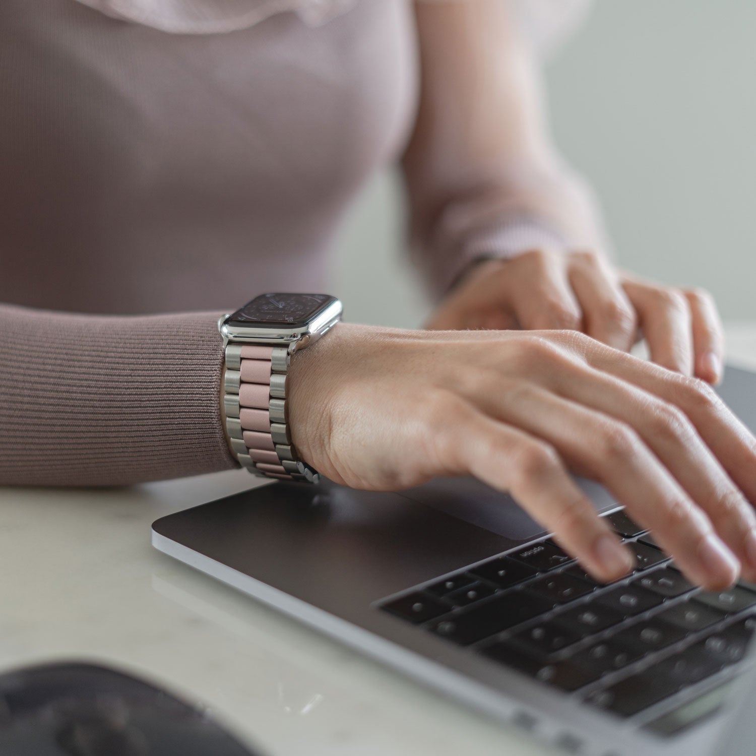 Eine Person in einem malvenfarbenen Langarmshirt tippt auf einem Laptop und trägt dabei das Arktisband Apple Watch Gliederarmband „Arctica“ im pink-silbernen Edelstahldesign.