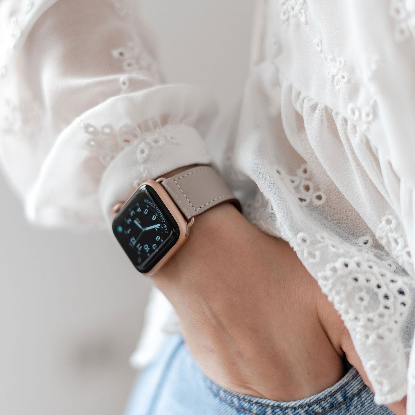 Nahaufnahme einer Person, die ein Arktisband Apple Watch Kunstlederarmband in Beige mit einer weißen bestickten Bluse trägt, Hand in der Tasche - ideal für maximalen Tragekomfort und einen minimalistischen Look.