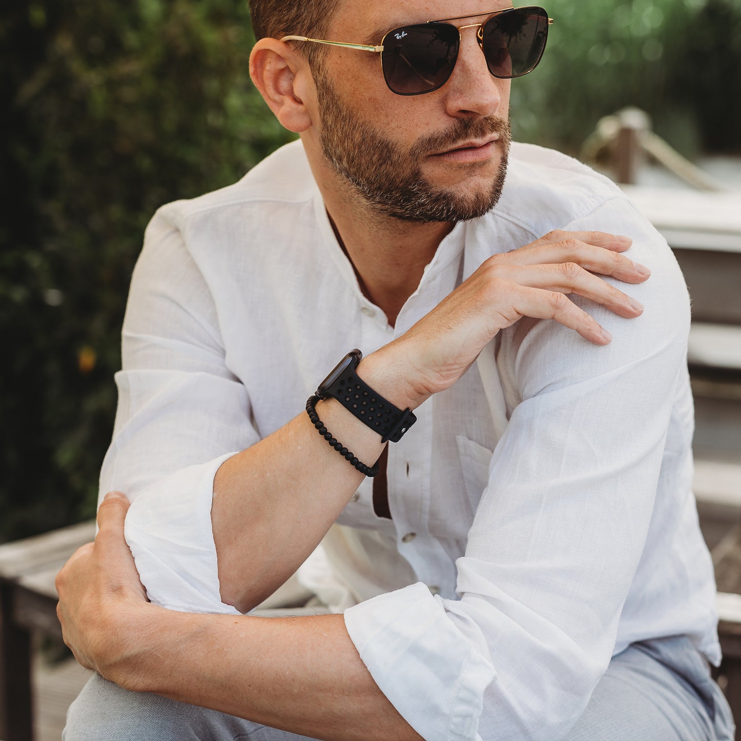 Ein Mann mit kurzen Haaren und Bart sitzt im Freien. Er trägt eine Sonnenbrille, ein weißes Hemd mit Knöpfen, ein Arktisband Apple Watch Silikonarmband „Energy“ und ein Perlenarmband. Er hat die Arme verschränkt und blickt zur Seite.