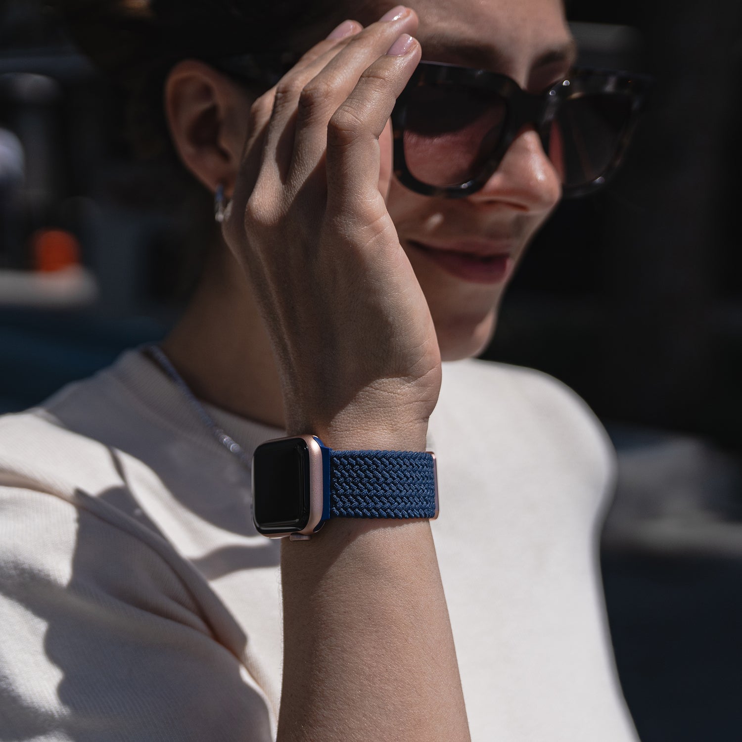 Frau im Freien mit Sonnenbrille und einer Apple Watch mit dem Arktisband Geflochtenes Apple Watch Armband „Casual“ mit Magnetverschluss in Blau, die ihr Gesicht berührt.