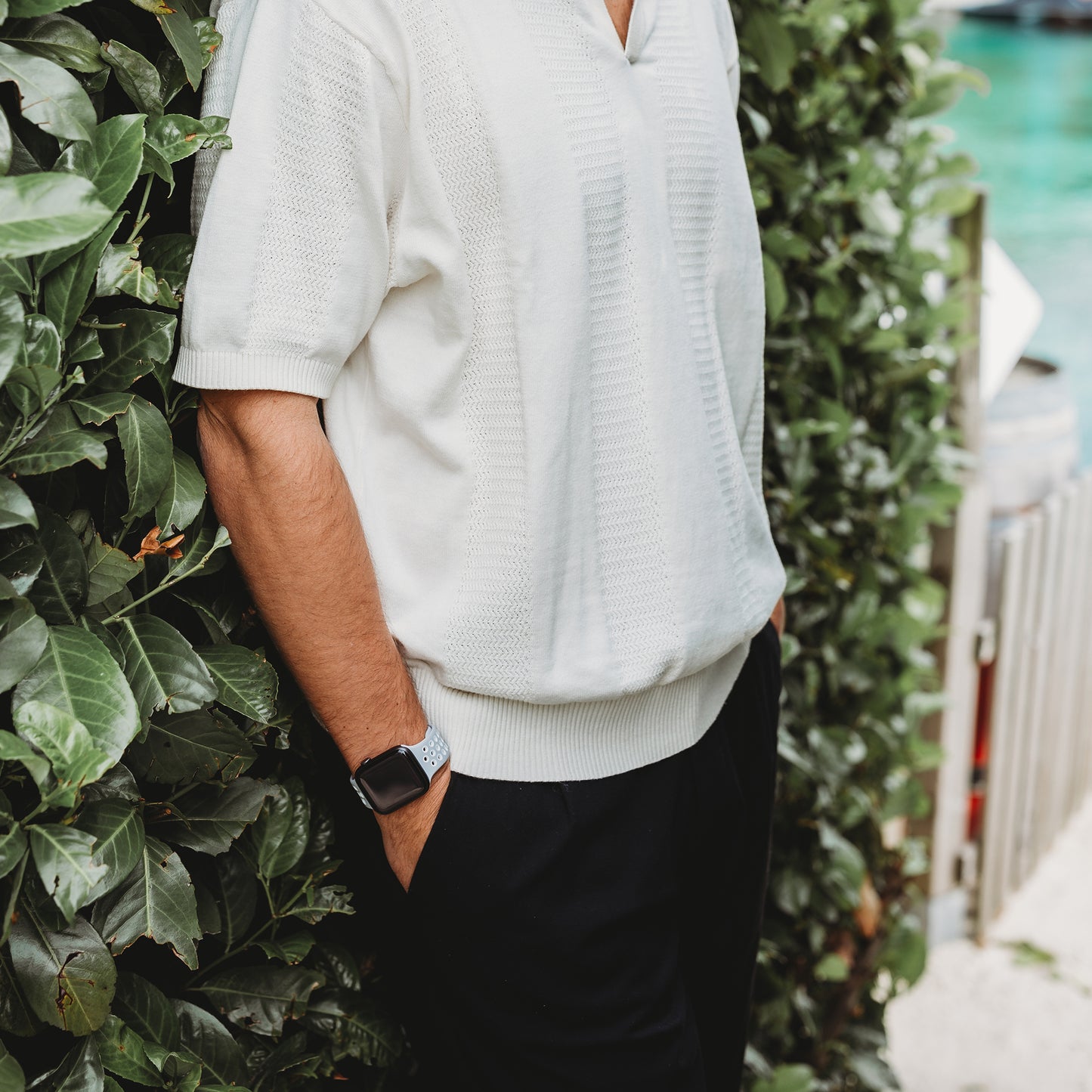 Eine Person in einem weißen kurzärmeligen Strickshirt, einer schwarzen Hose und dem arktisband Apple Watch Silikonarmband „Energy“ von arktisband steht mit den Händen in den Taschen neben einer grünen Hecke, im Hintergrund sind Wasser und ein Zaun zu sehen.