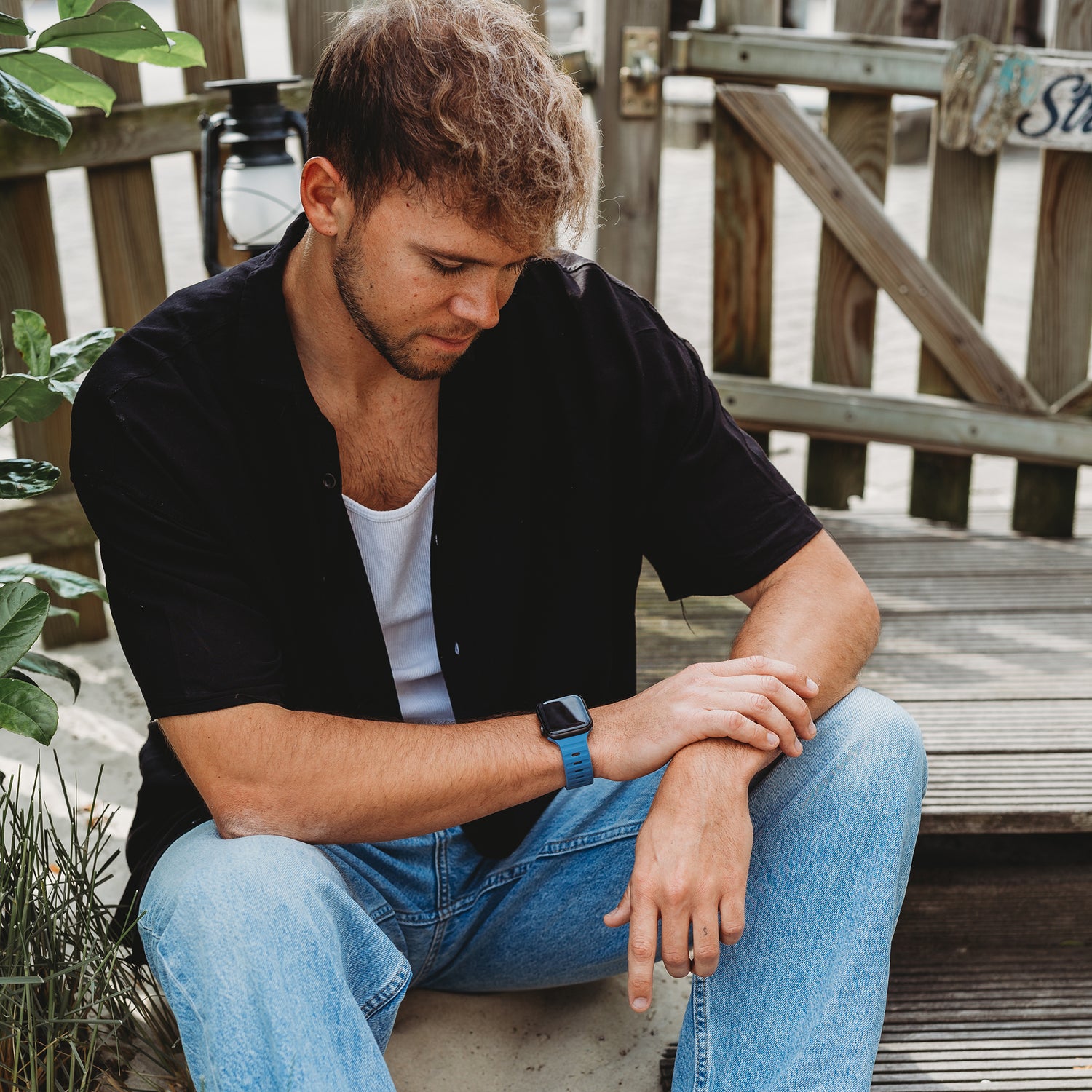 Ein Mann mit hellbraunem Haar, schwarzem Hemd, weißem T-Shirt und Blue Jeans sitzt im Freien auf Holzstufen, trägt eine Apple Watch Ultra mit dem arktisband „Active“ Silikonarmband von arktisband und blickt nach unten, sein Arm ruht auf seinem Knie.