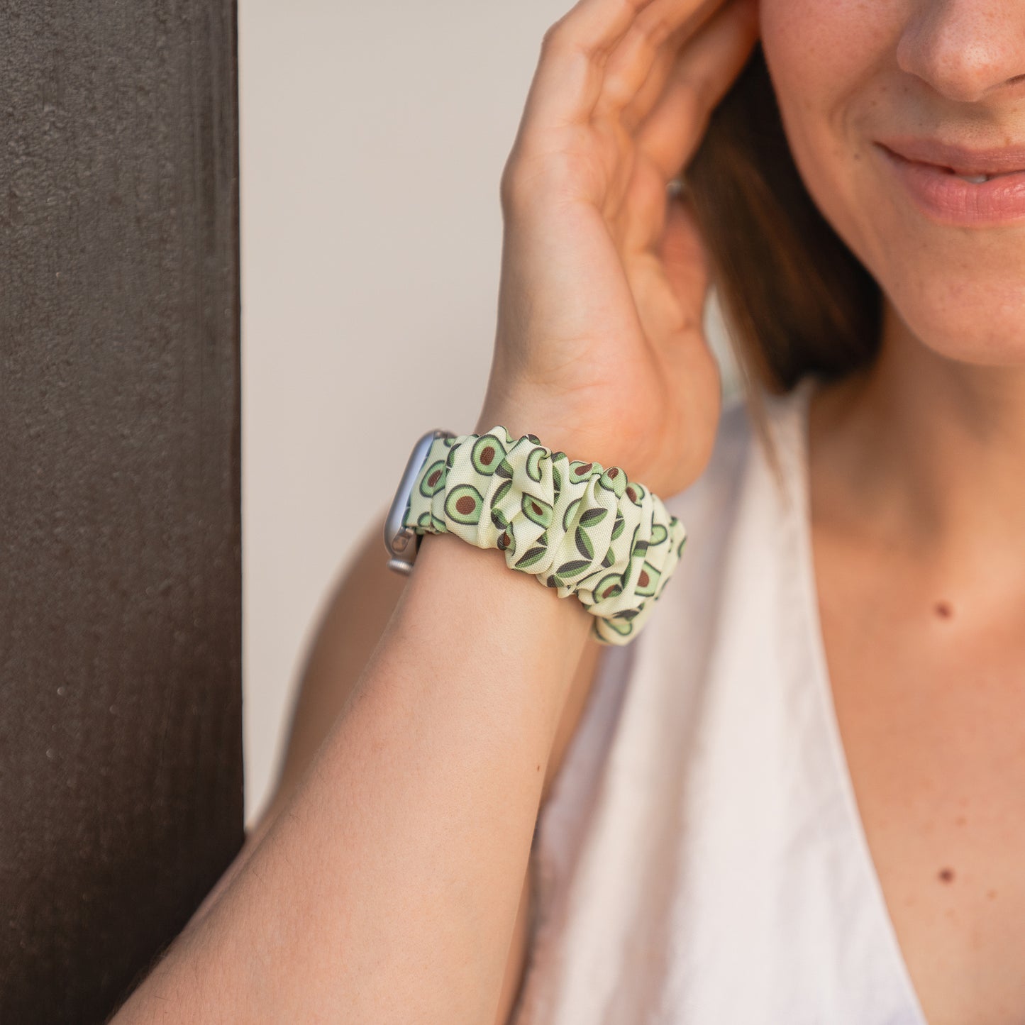 Eine Frau lächelt und berührt ihr Gesicht, während sie das arktisband Apple Watch Scrunchie Armband "Elanora" mit einem Avocado-Muster auf ihrer Smartwatch trägt.