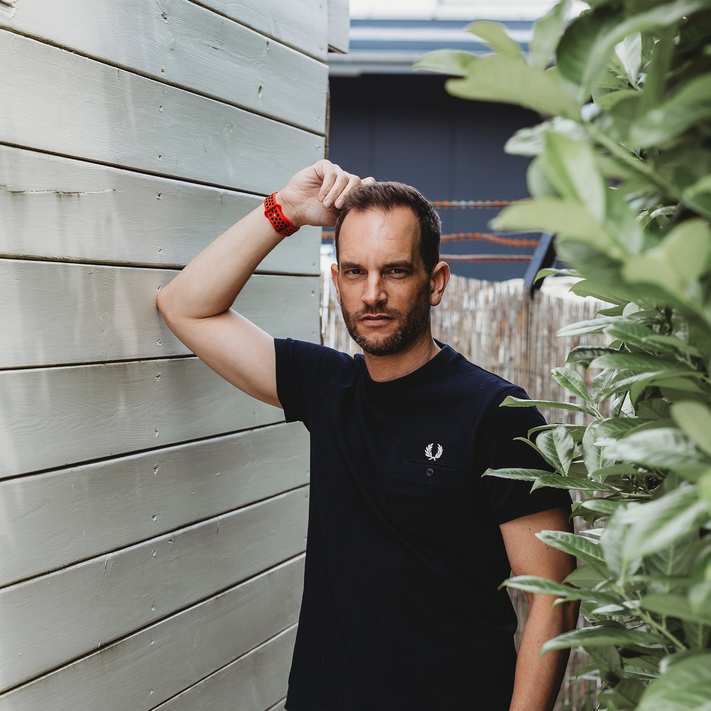 Ein Mann in einem schwarzen T-Shirt und einem Arktisband Apple Watch Silikonarmband „Energy“ steht im Freien an einer hellen Holzwand und grünen Pflanzen, stützt seinen Arm auf die Wand und blickt in die Kamera.