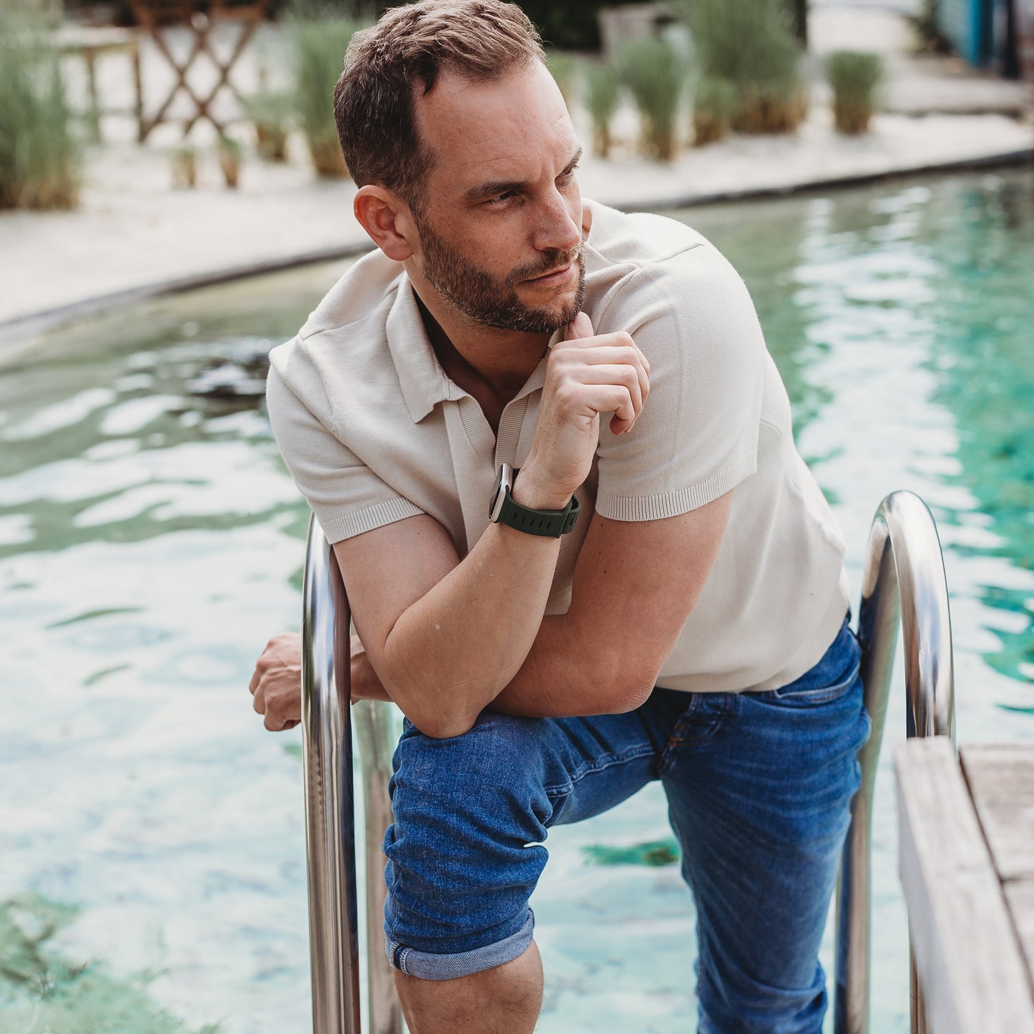 Ein Mann mit kurzen Haaren und Bart, bekleidet mit einem hellen Poloshirt und hochgekrempelten Jeans, lehnt an einer metallenen Poolleiter und trägt das arktisband Apple Watch Silikonarmband „Active“. Im Hintergrund sind grüne Pflanzen und Gartenmöbel zu sehen.