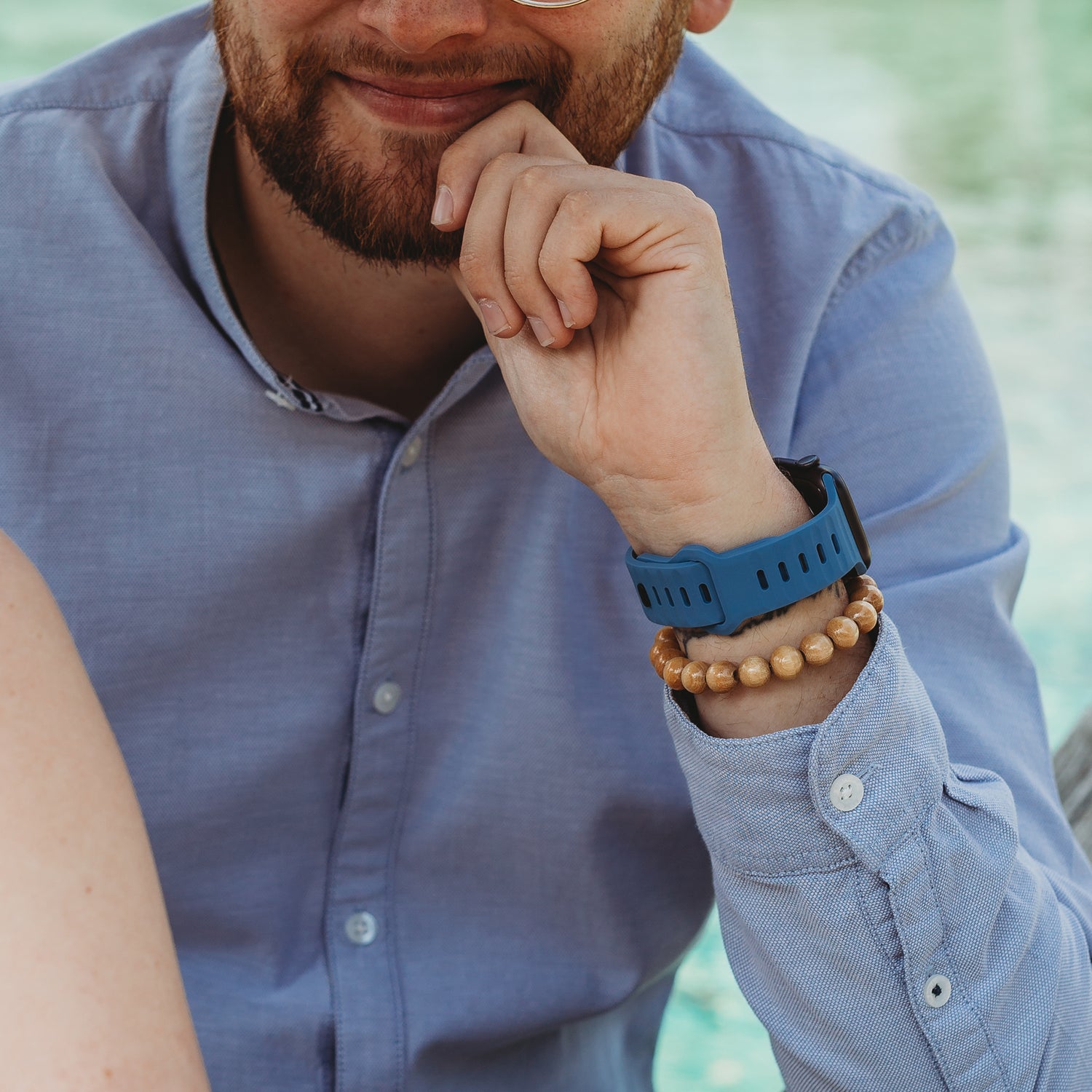Eine Person in einem hellblauen Hemd trägt ein Arktisband Apple Watch Silikonarmband „Active“ und Holzperlenarmbänder und stützt ihr Kinn auf die Hand. Der unscharfe Hintergrund lässt auf eine Außenkulisse am Wasser schließen.