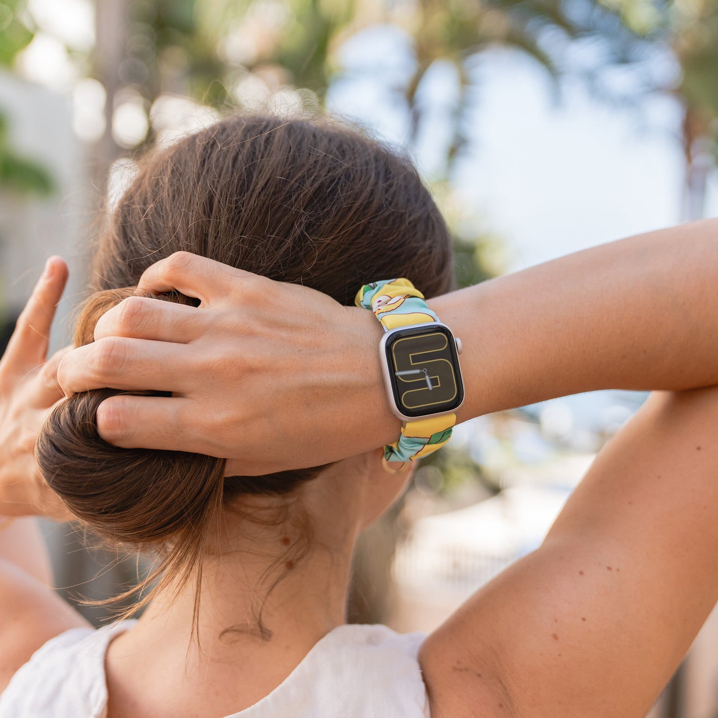 Eine Frau mit hochgestecktem Haar trägt das arktisband Apple Watch Scrunchie Armband "Elanora" von arktisband, das durch sein farbenfrohes, elastisches Design besticht, während sie sich im Freien mit den Händen hinter dem Kopf entspannt.